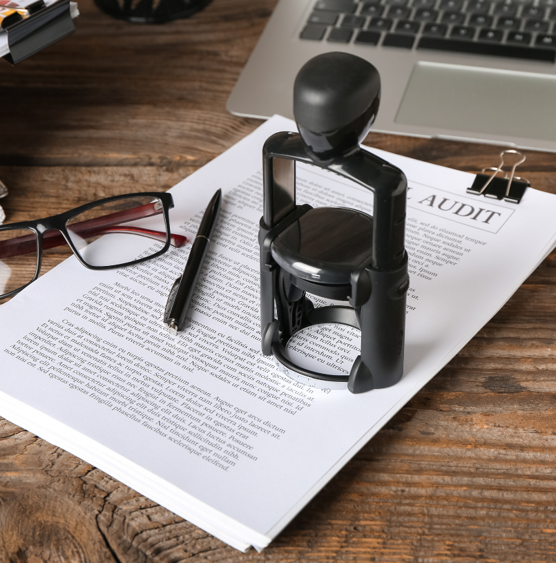 Office desk with glasses, a pen, a stamp, a document labeled 'AUDIT,' a laptop, and a binder clip on a wooden surface.