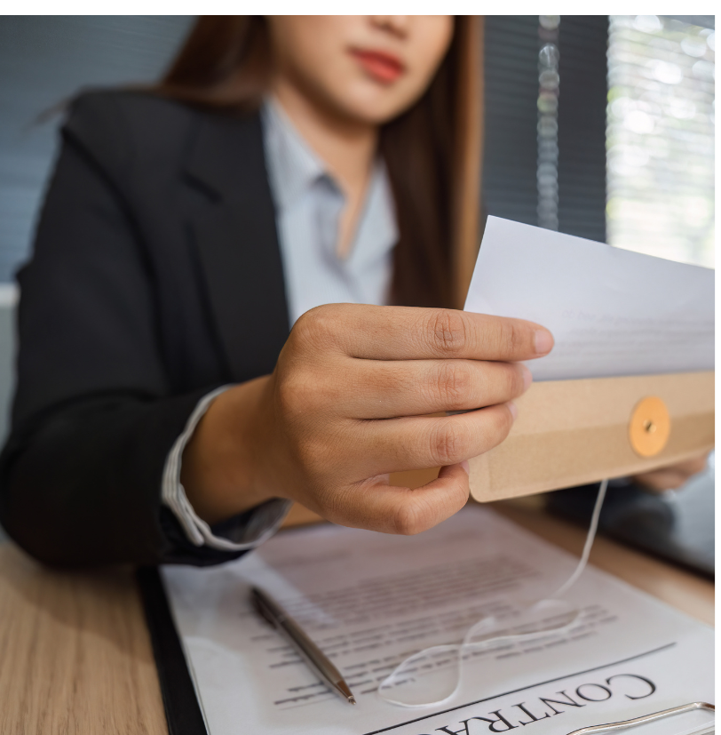 A woman in business attire holding a piece of paper or document in her hand, sitting at a desk with a pen and a clipboard.