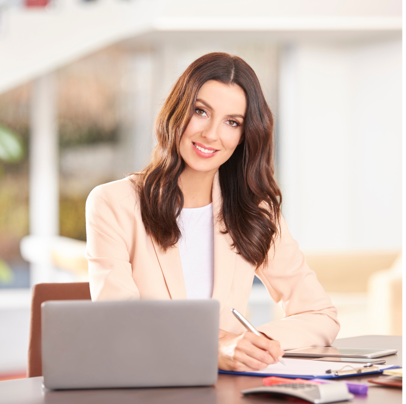 Businesswoman sitting at a desk with a laptop, smiling and holding a pen, in a bright office space.