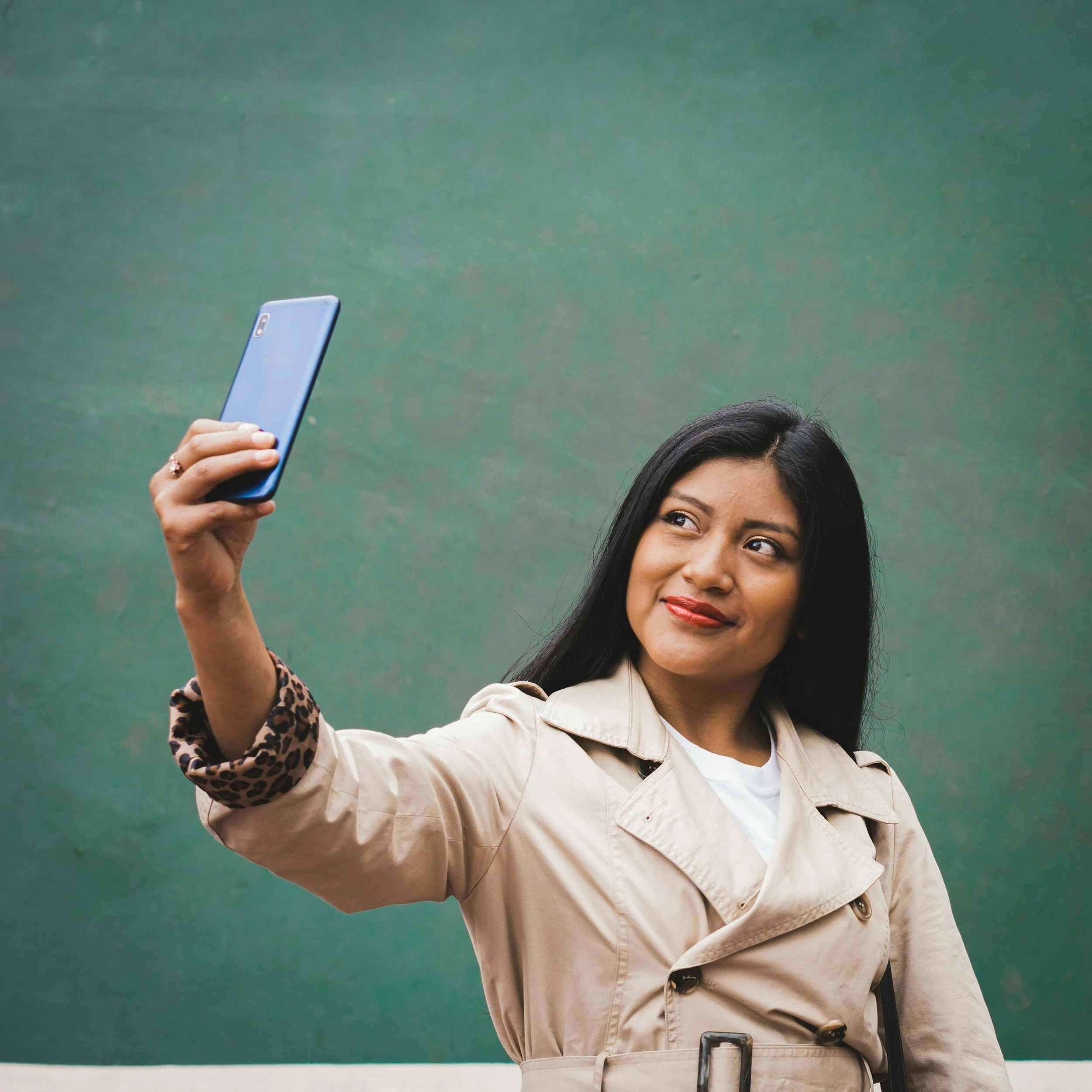 Woman with long black hair taking a selfie with a blue smartphone against a plain green background.