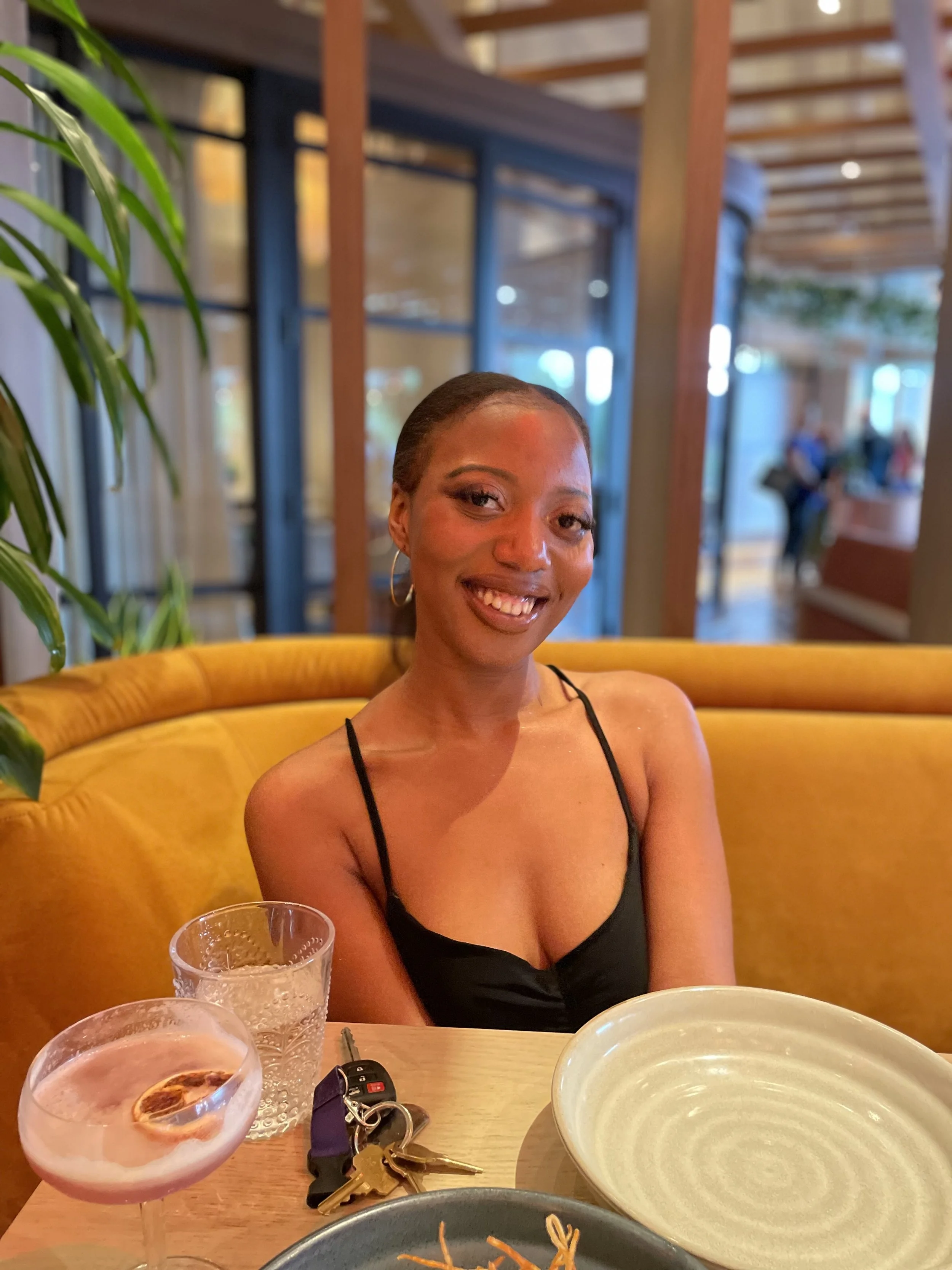 A woman with dark hair seated at a restaurant table smiling, with a yellow booth behind her, a glass of pink drink with a lemon slice, a glass of water, keys, and an empty white bowl on the table, and plant greenery on the side.