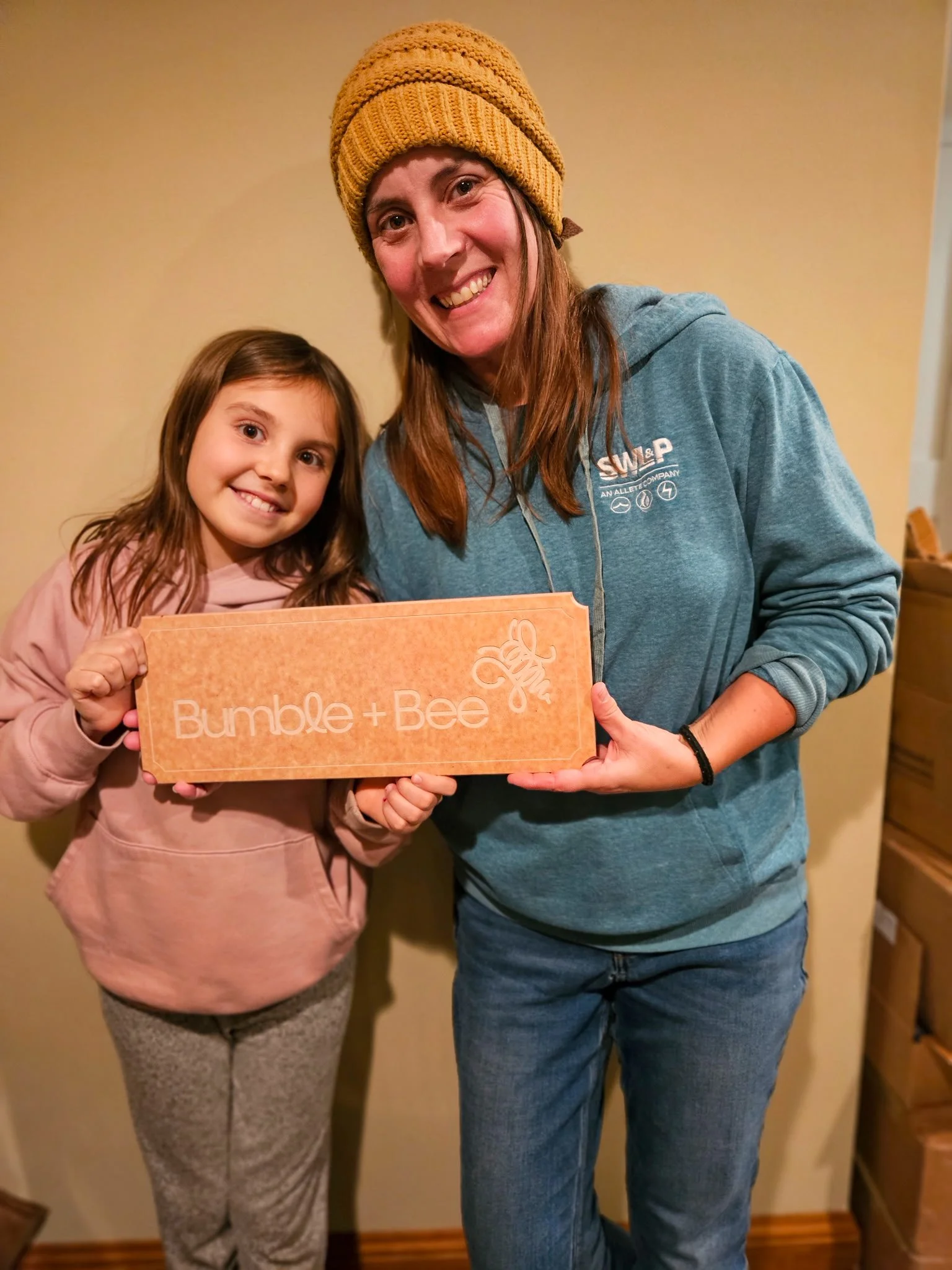 A mother and a daughter holding a sign that says 'Bumble + Bee' standing indoors against a beige wall.