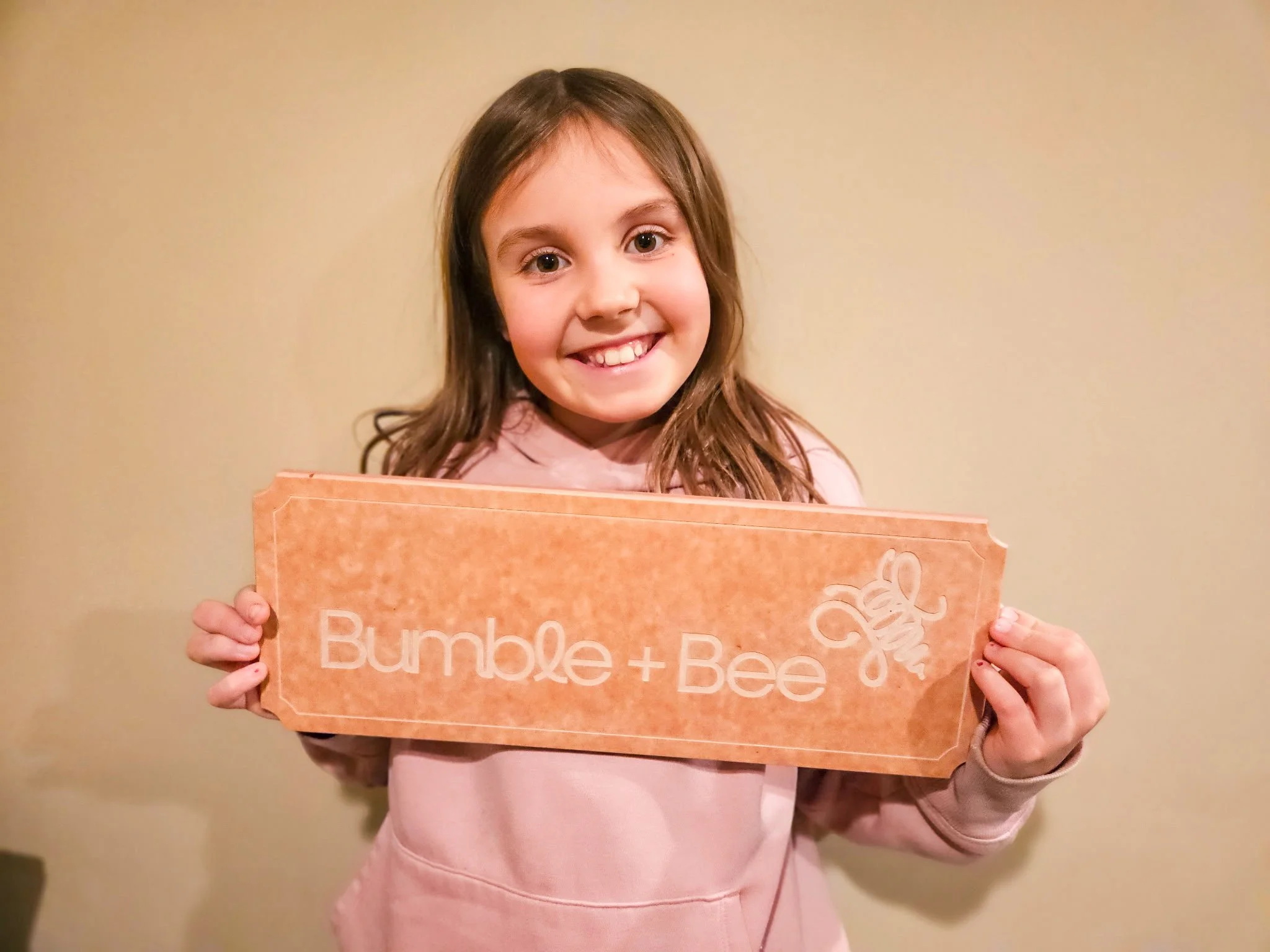 A smiling young girl with brown hair holding a sign that says "Bumble + Bee" with a drawing of a bee.