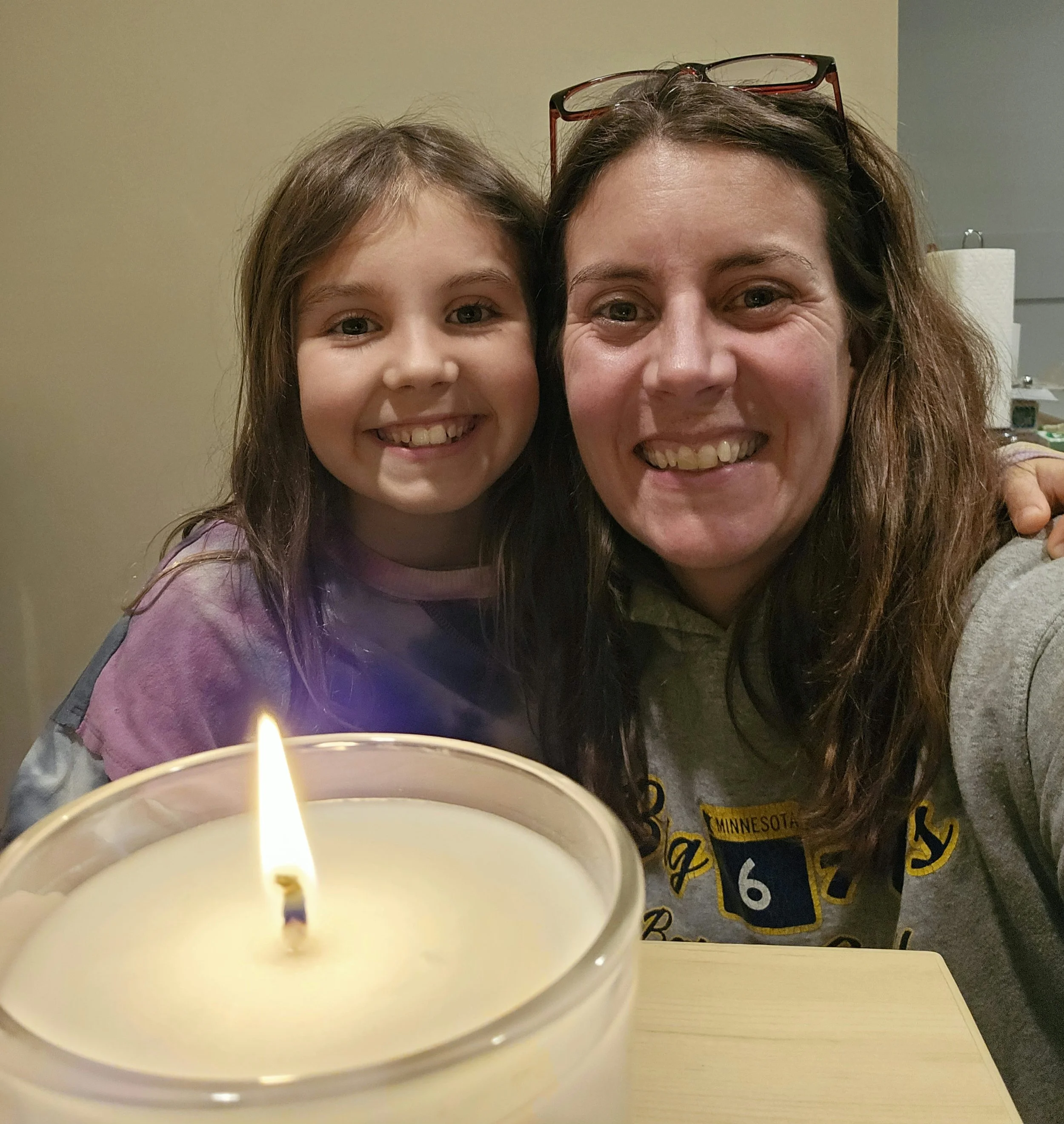 A mother and her young daughter smiling at the camera while sitting at a table with a lit candle in the foreground.