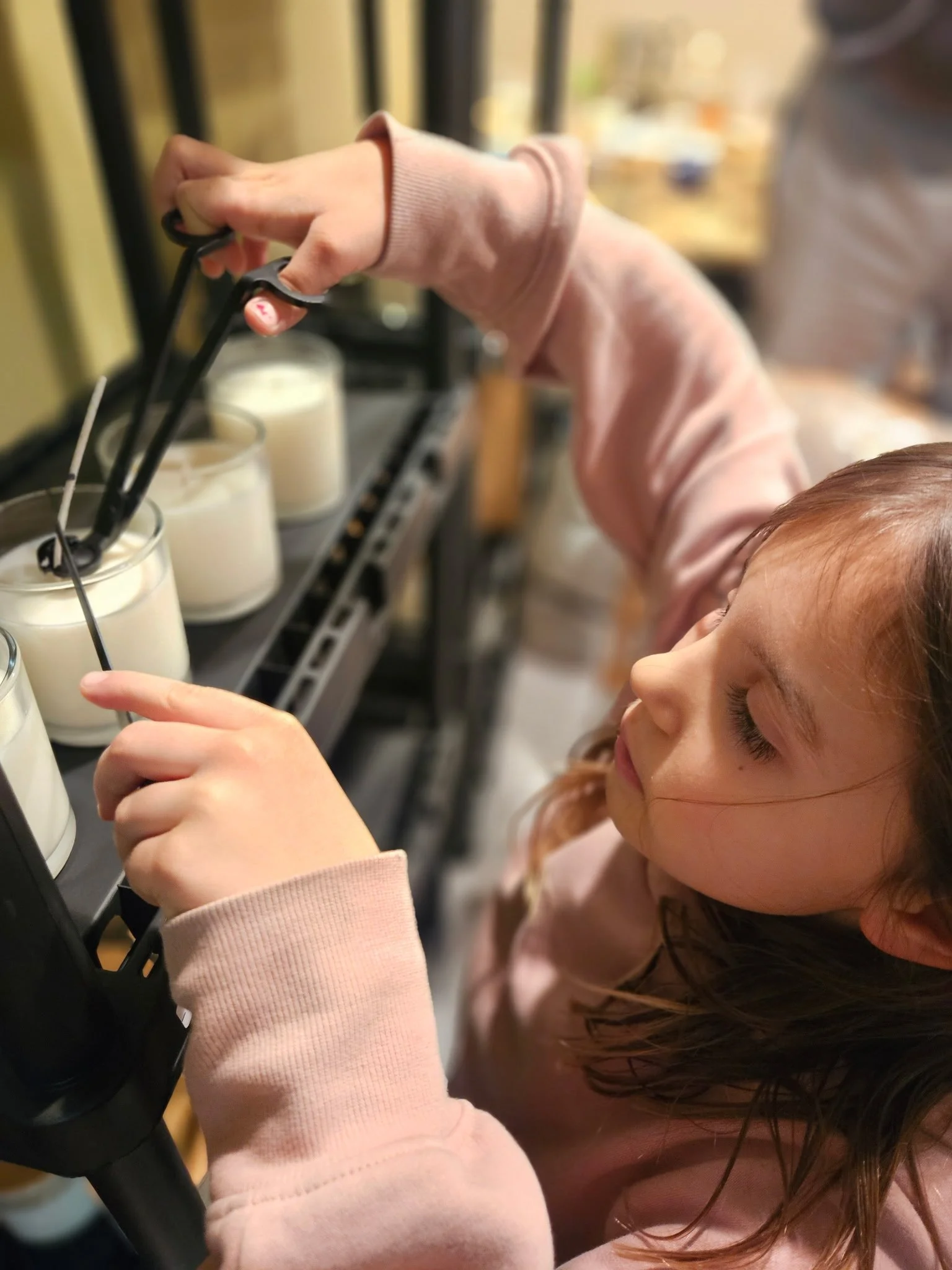 A young girl with long brown hair is placing a candle into a glass container using tongs in a store.