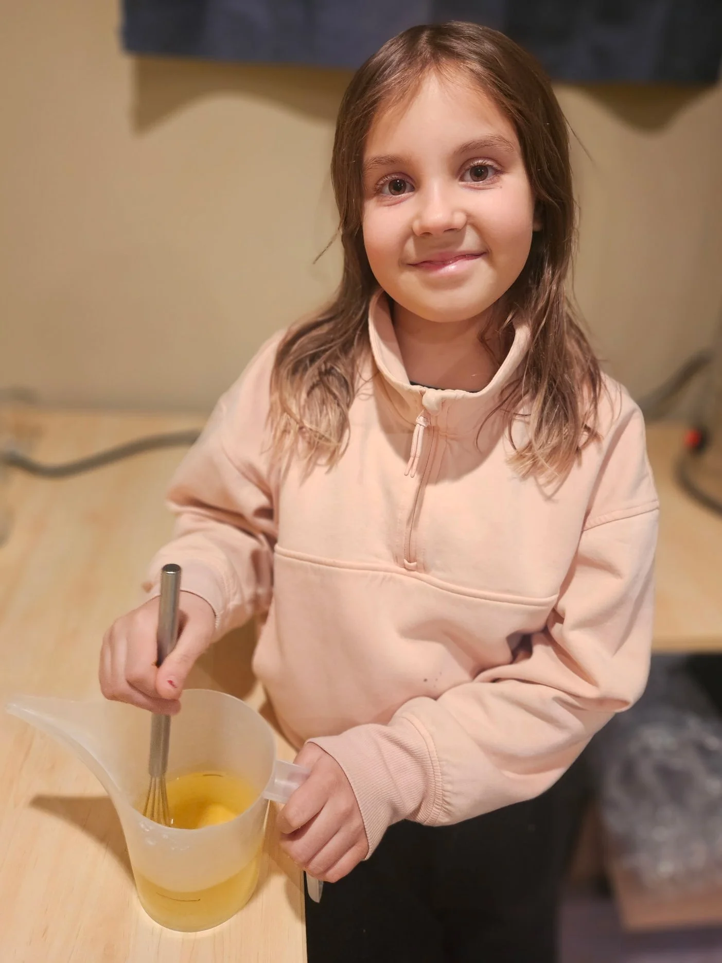 A young girl with long brown hair and light skin smiling at the camera while whisking yellow liquid in a clear plastic measuring cup.