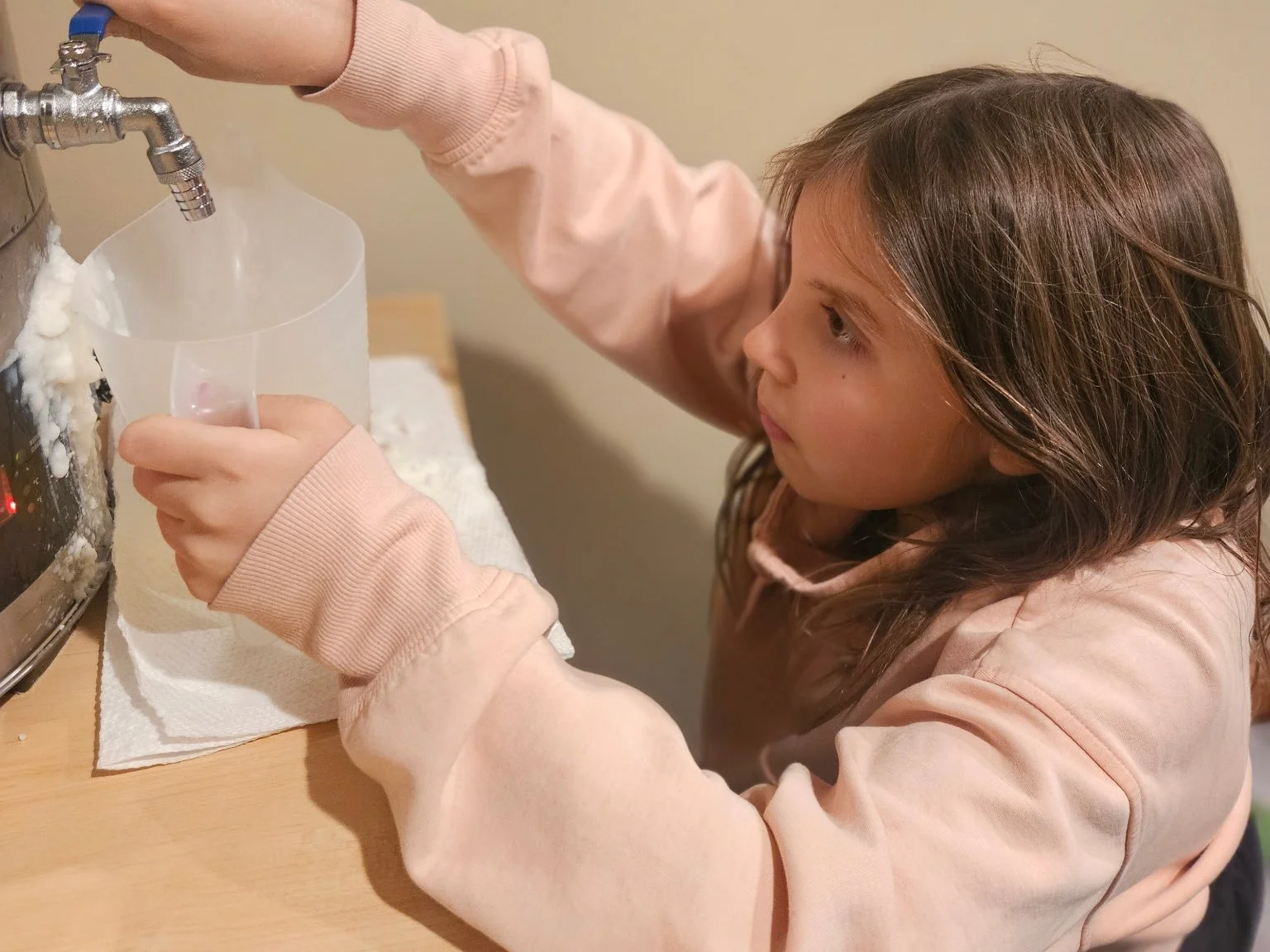 A young girl with brown hair wearing a pink sweatshirt pouring a liquid from a faucet into a clear plastic cup over a paper towel on a wooden table.