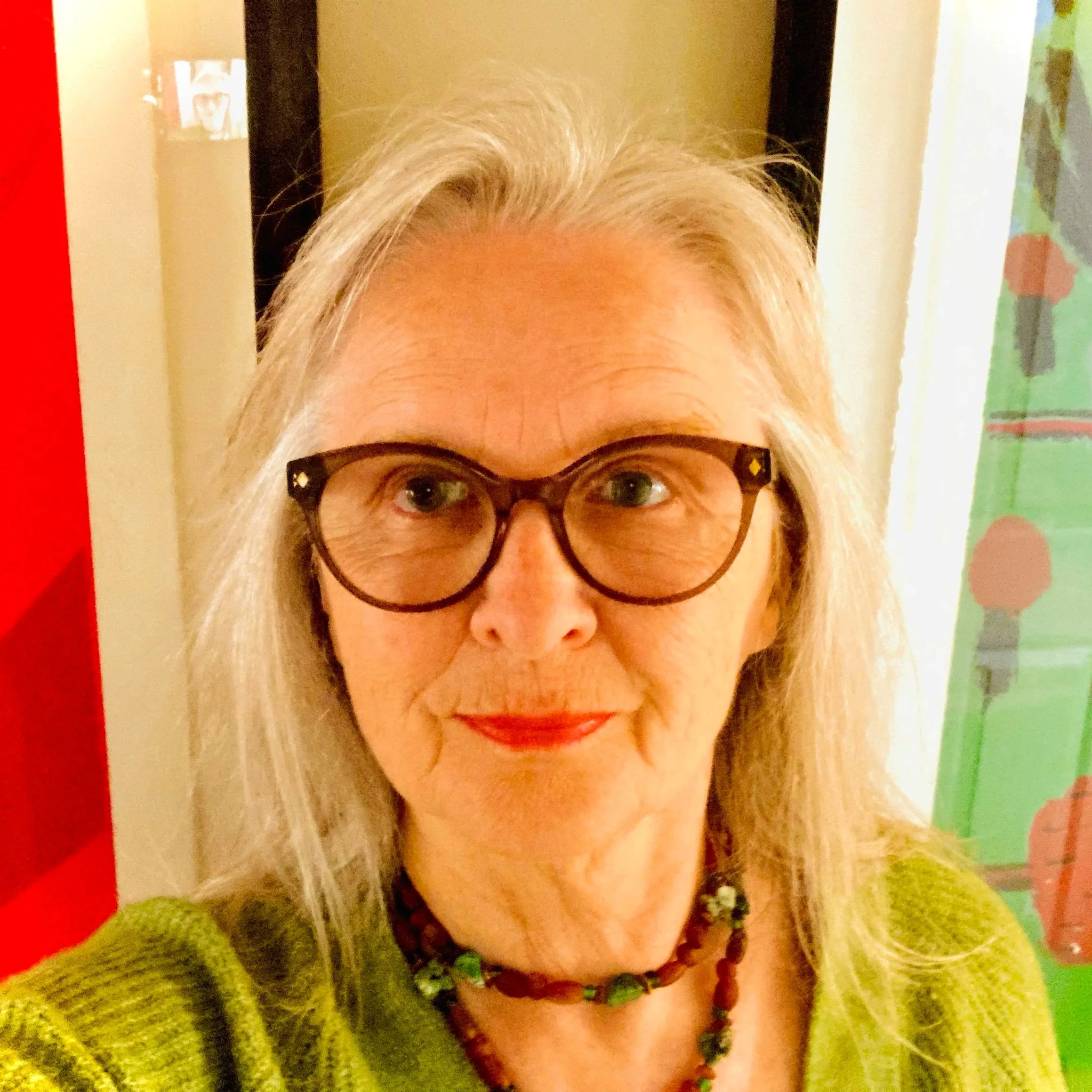 A woman with long gray hair wearing glasses, a green sweater, and a beaded necklace, smiling at the camera indoors.