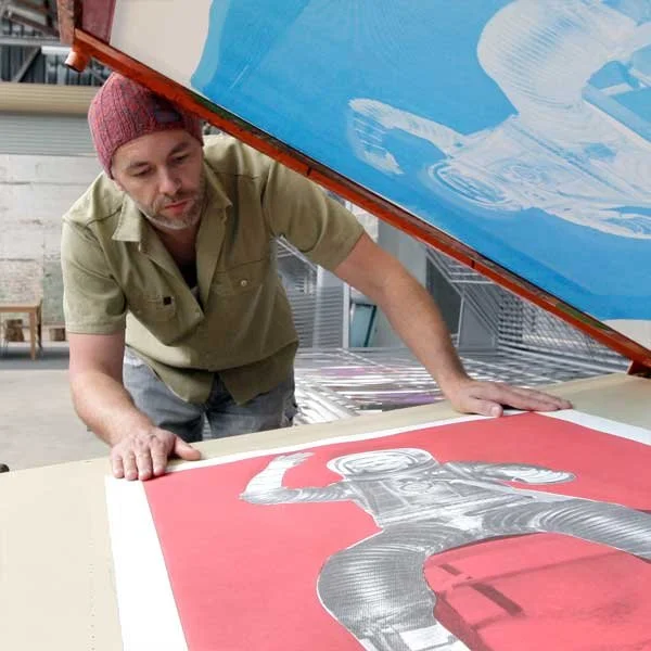 An artist is working on a large canvas with a red background, depicting a figure in black and white. The artist is wearing a beige shirt and a red beanie, leaning over the artwork in a studio setting.