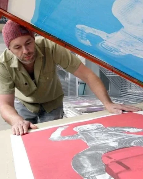 Nicholas Mau working on a large pop art style print of Richard Branson. He is leaning over the print while adjusting or inspecting it on a flat surface.