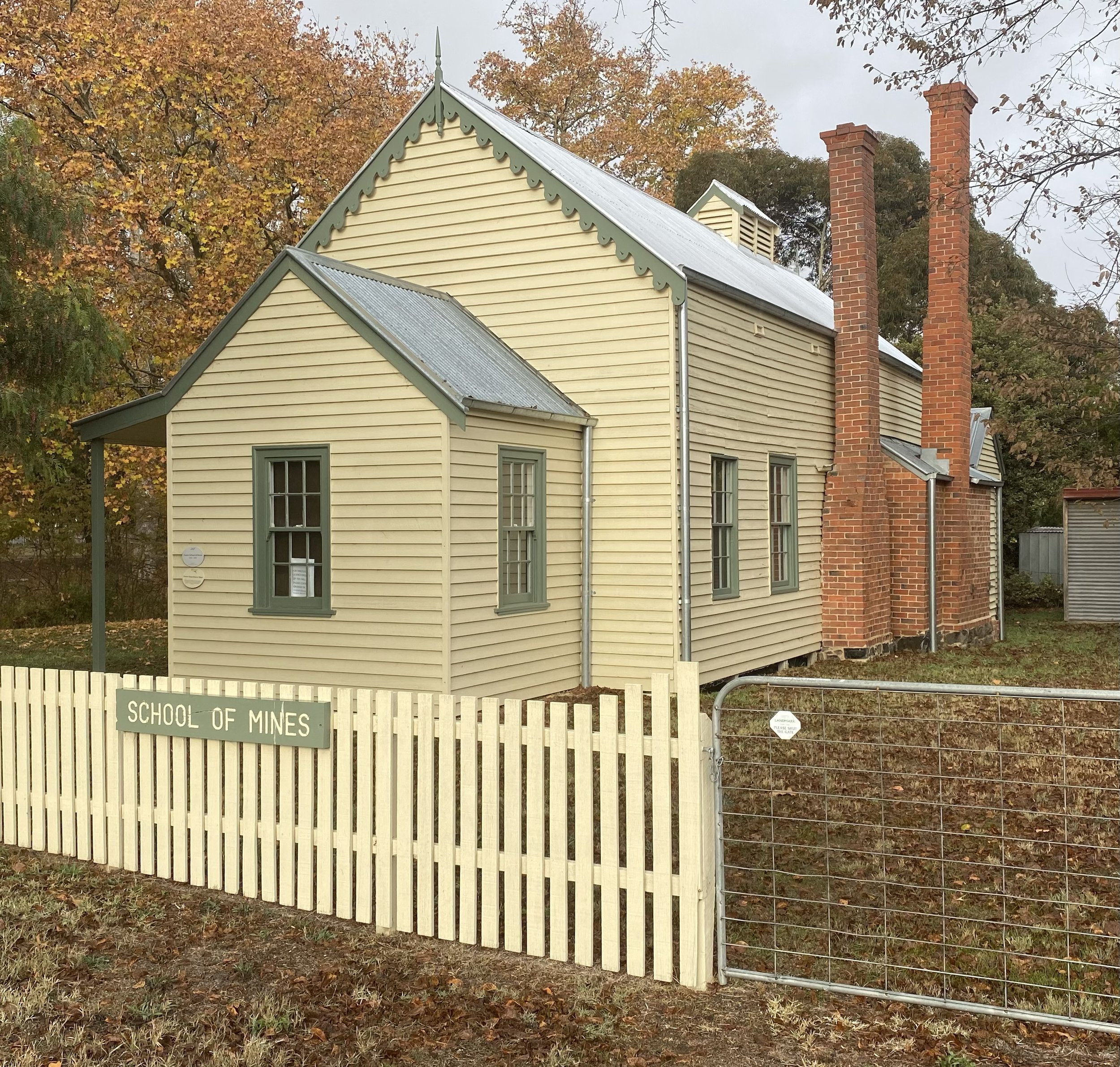 A historic yellow wooden school building with green trim and a metal roof, surrounded by a cream picket fence with a sign that reads 'School of Mines.' The building has multiple windows and two tall brick chimneys, set against a backdrop of autumn trees.