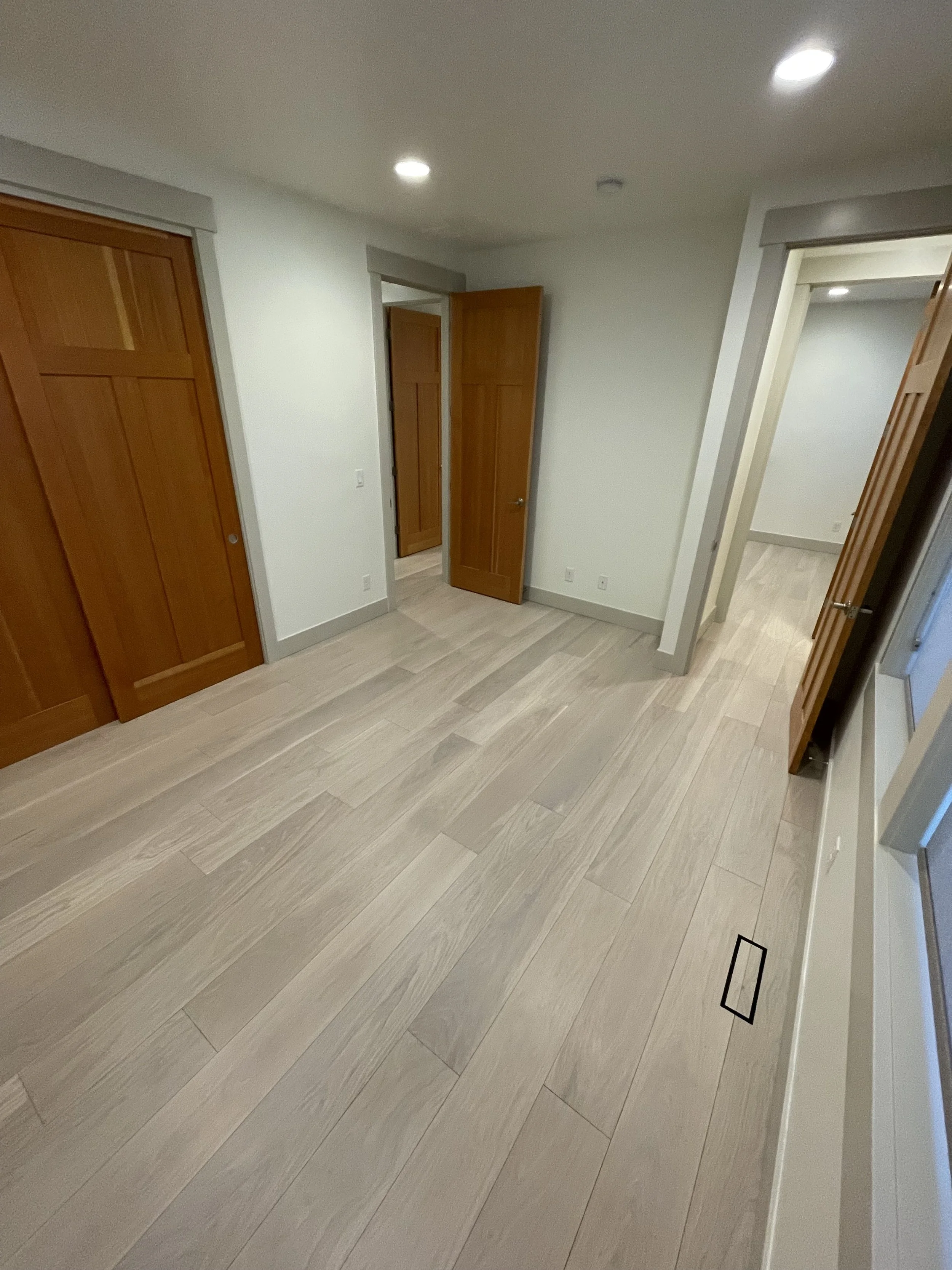 Empty room with white walls, light wood flooring, and wooden closet doors. Two recessed ceiling lights are visible, and a small window or vent near the floor.