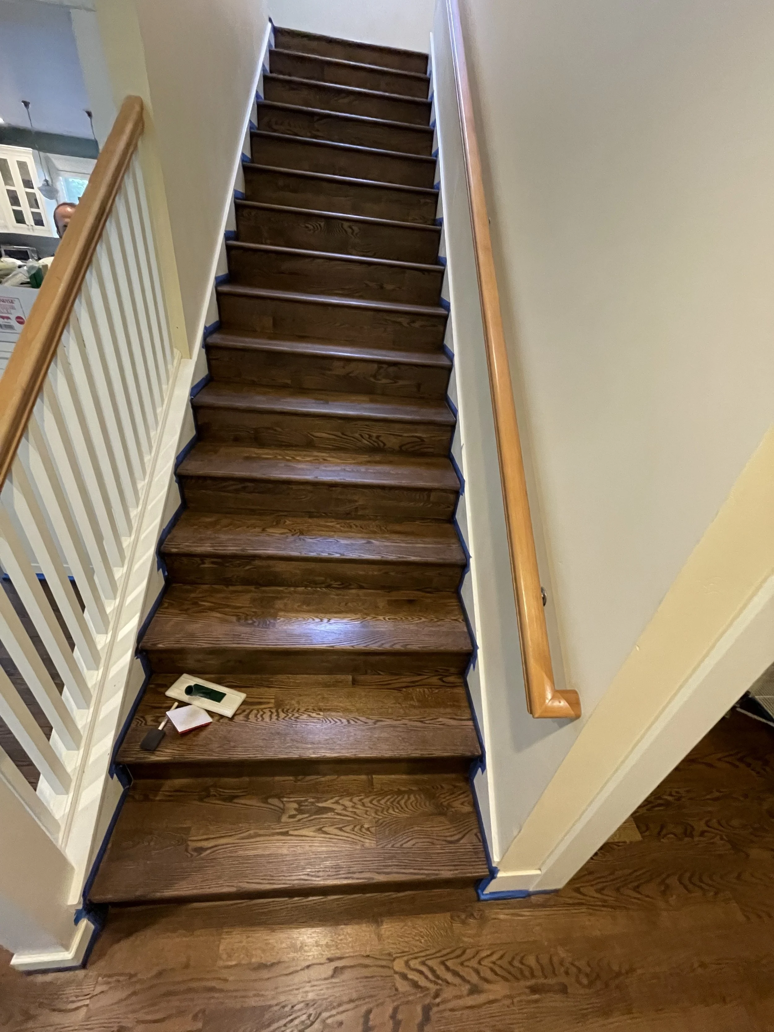 Inside view of a staircase with wooden steps being refinished, with tools and tape on the first step, and a wooden handrail on the right side.