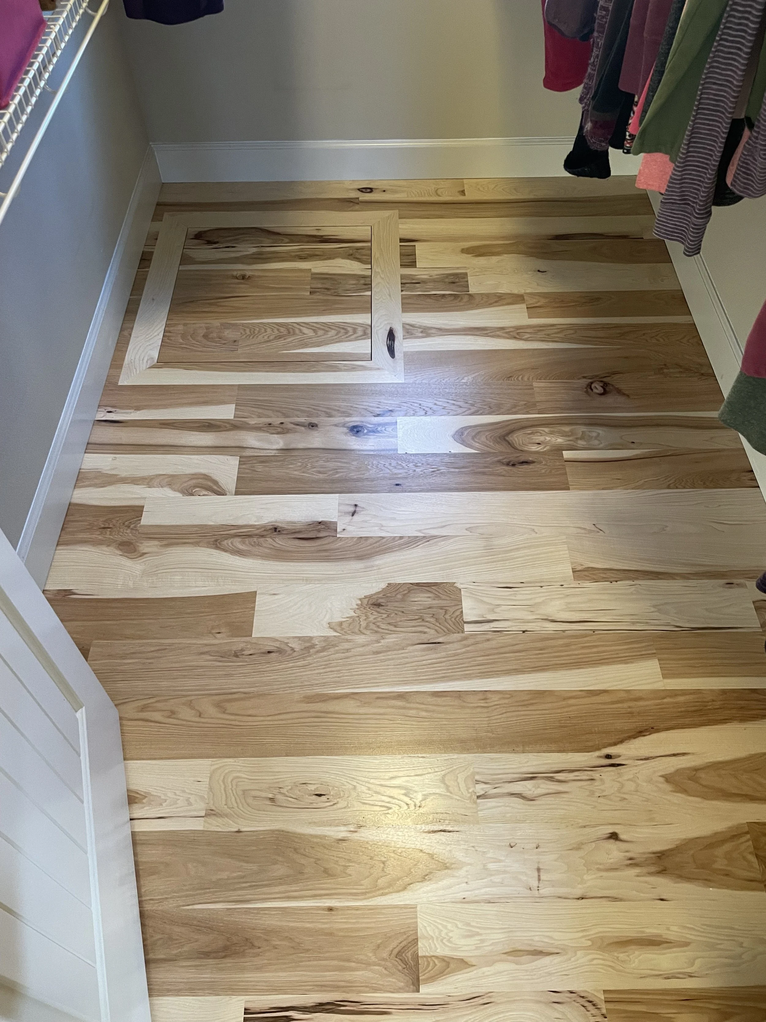 Wooden floor with a small access panel near the back wall, surrounded by hanging clothes on the right side and a white wire shelf on the left side.