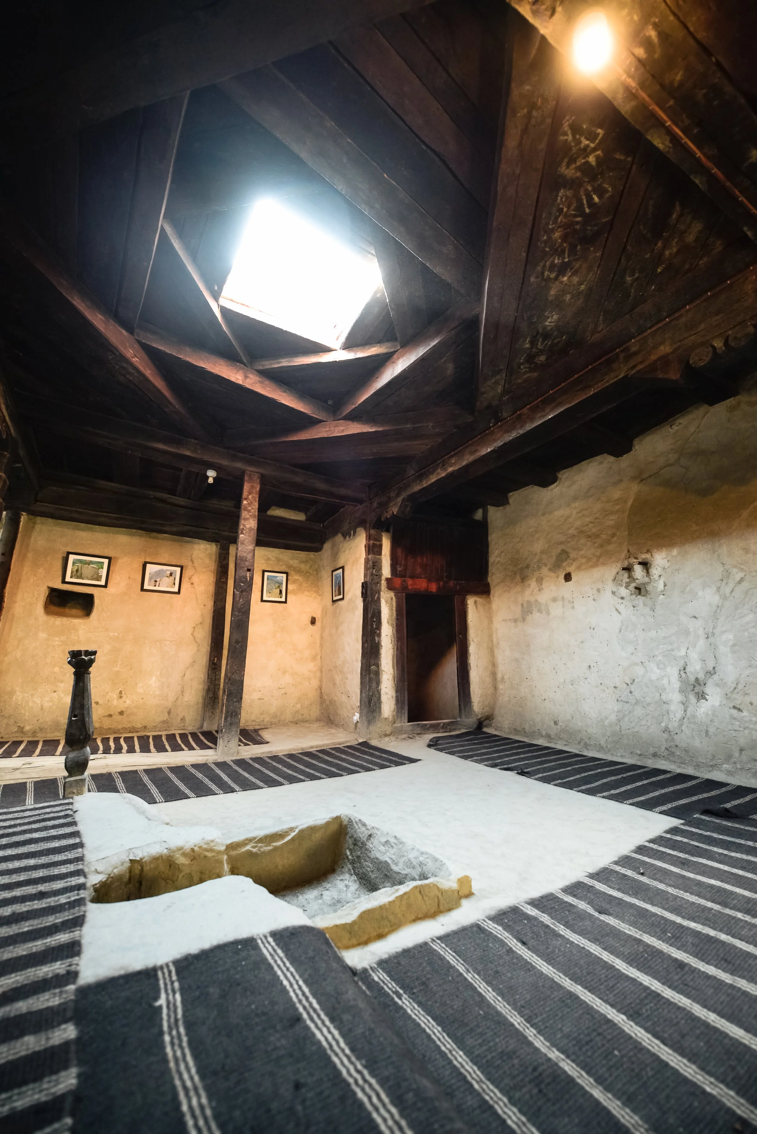 Interior of a rustic room with a skylight in the ceiling, wooden beams, framed pictures on the wall, and a hole in the floor surrounded by striped rugs.