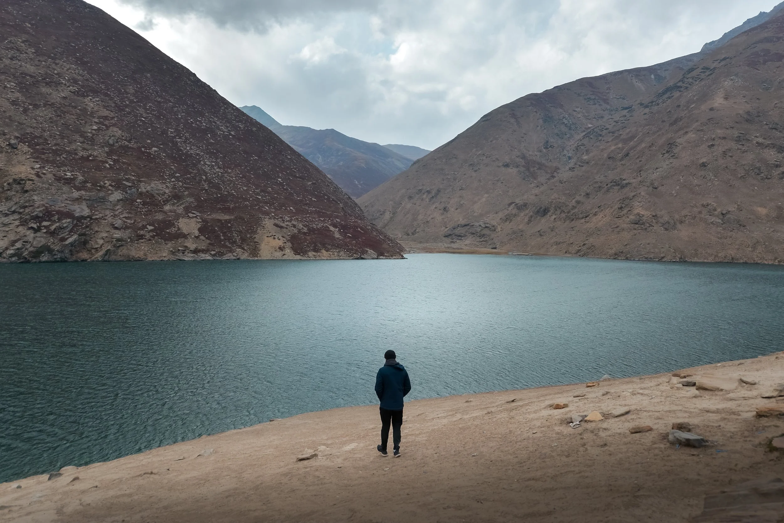 A person in a blue jacket and black pants walking on a sandy shore towards a calm mountain lake, surrounded by brown and gray mountains under cloudy skies.