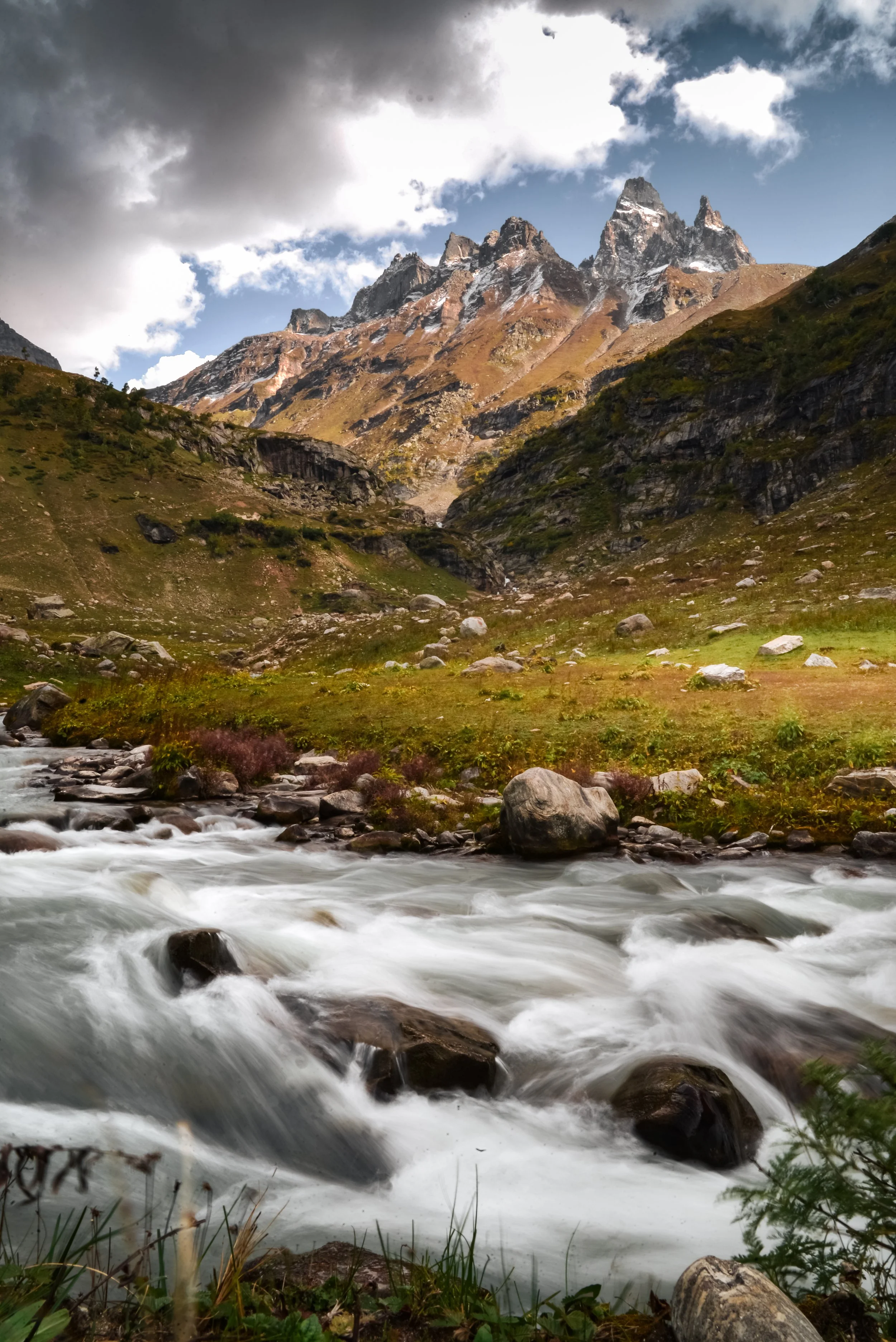 Mountain landscape with rushing river, rocky terrain, green meadows, and rugged peaks with snow, under cloudy sky.