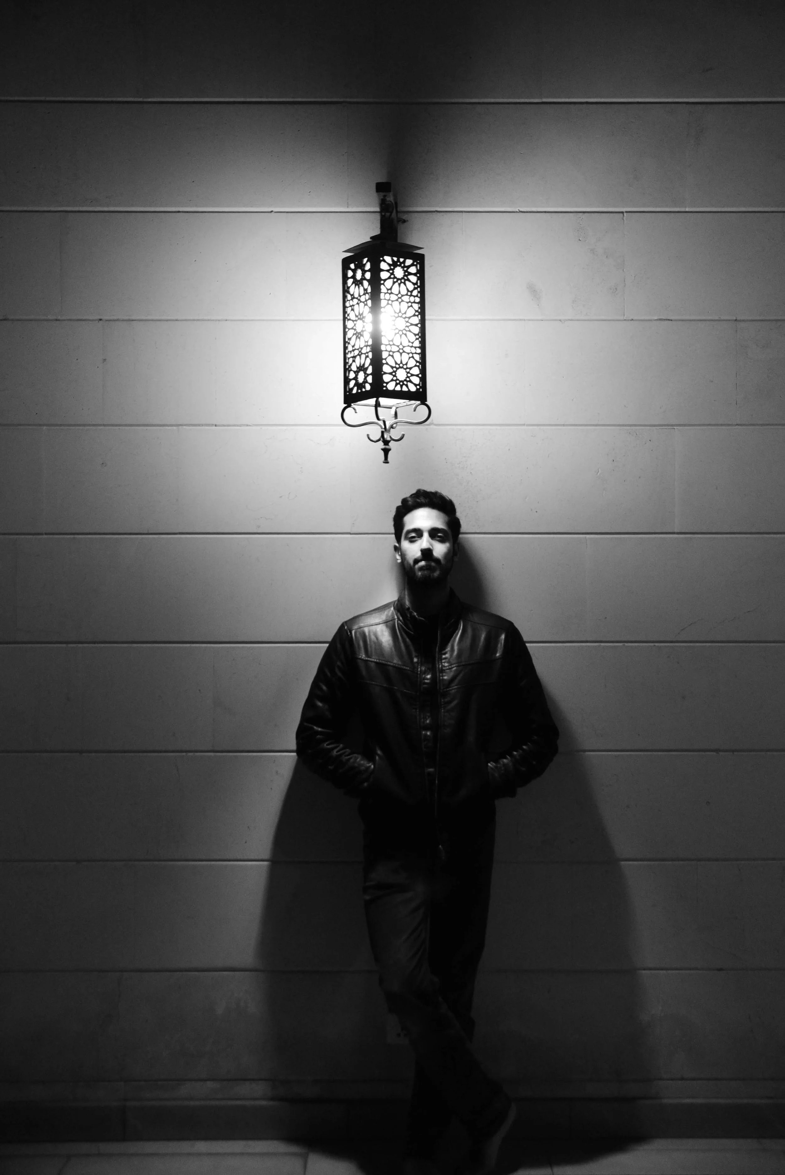 A man dressed in a leather jacket standing against a wall with his hands in his pockets, illuminated by a hanging lantern above him, in black and white.