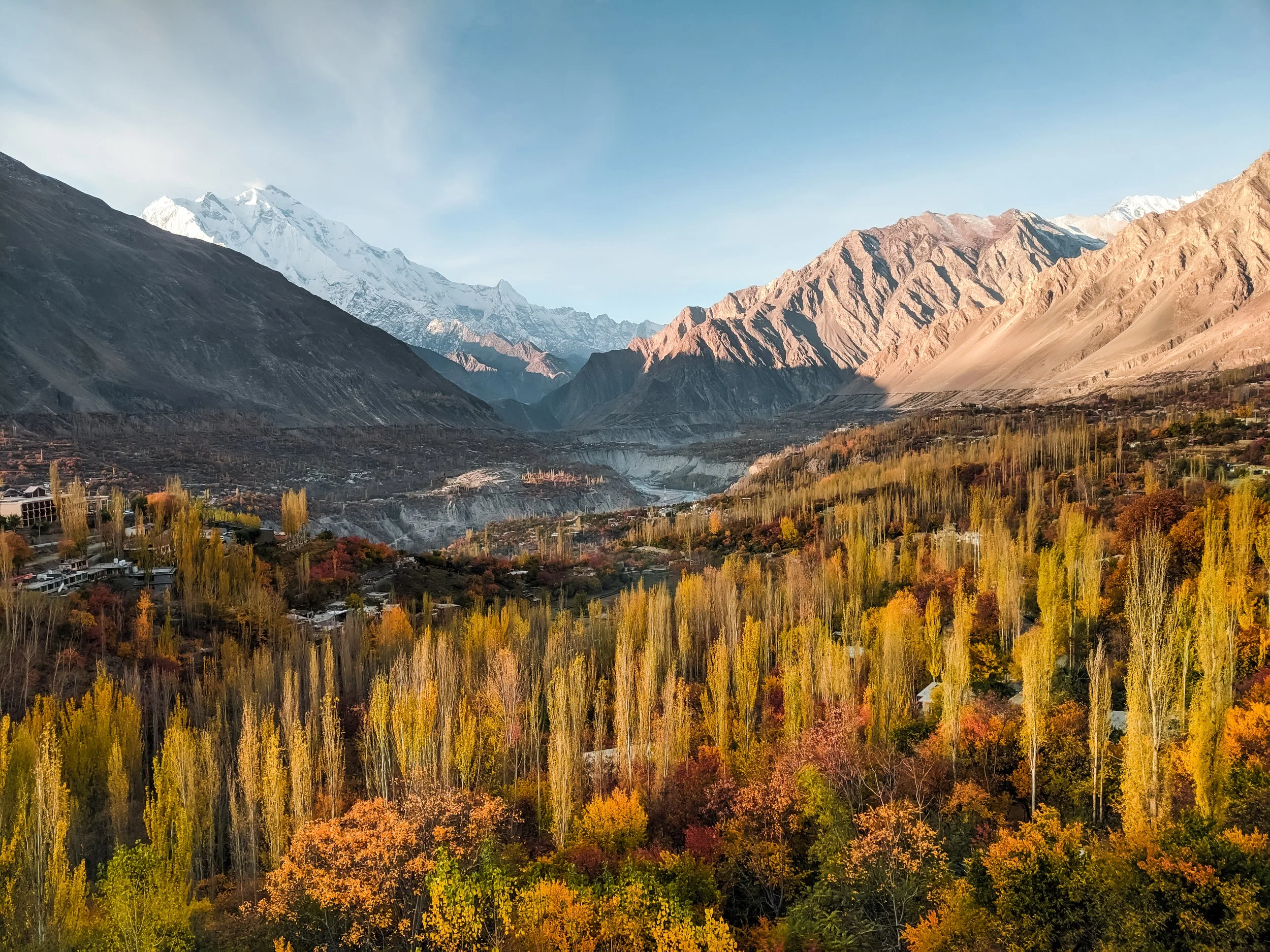 Scenic landscape of a valley with colorful autumn trees, mountains with snow-capped peaks in the background, and a lake in the middle distance.