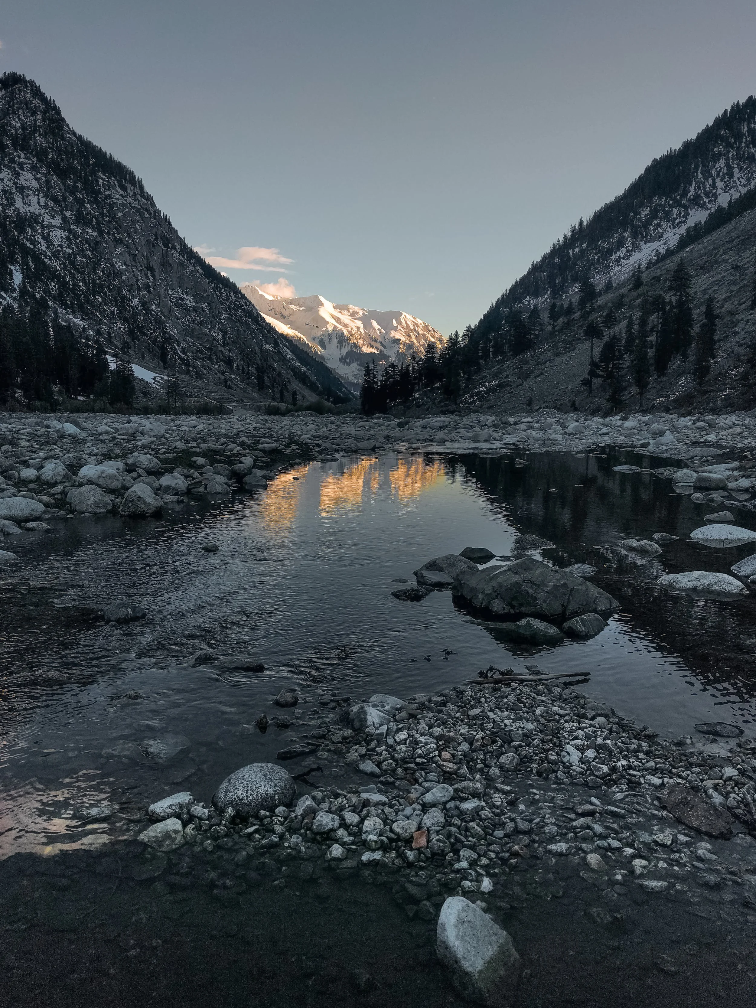 A mountain river flowing through a rocky valley with snow-capped mountains in the background under a clear evening sky.