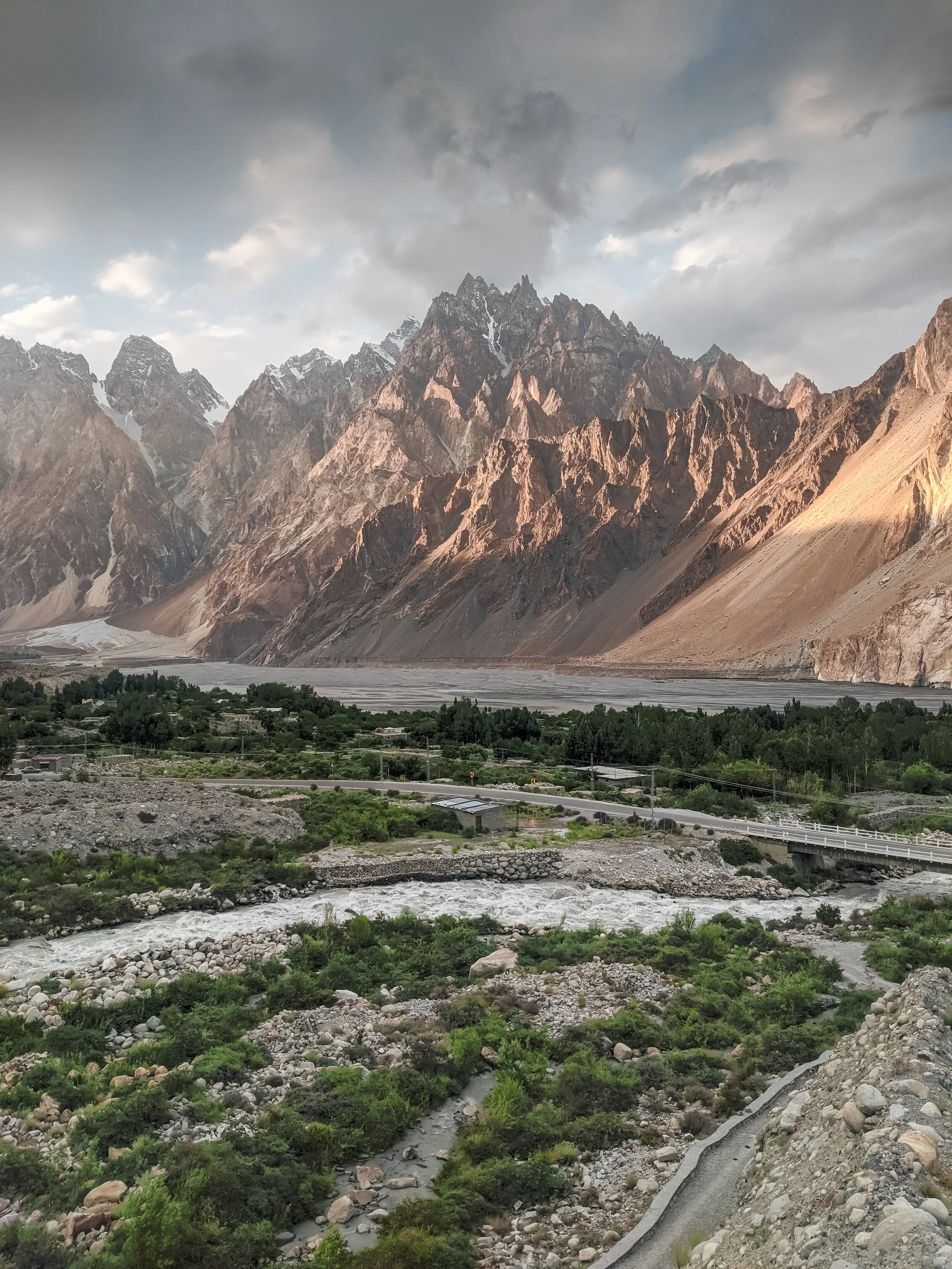 A mountain landscape with steep rocky peaks, a river at the base, green vegetation, and a cloudy sky.