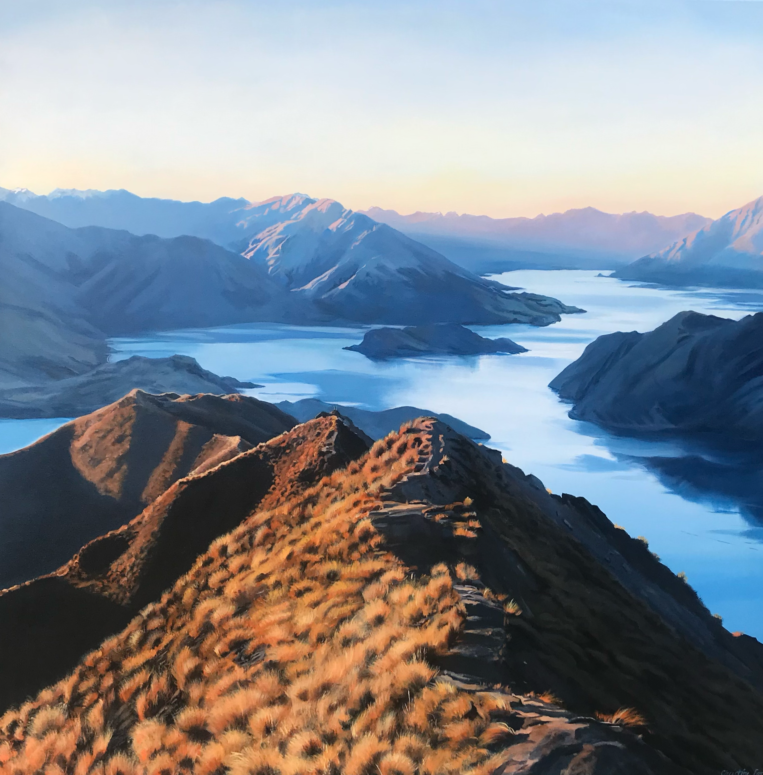 Landscape view of a mountain range with water bodies, showing snowy peaks and a ridge covered with orange and yellow grass during sunset.