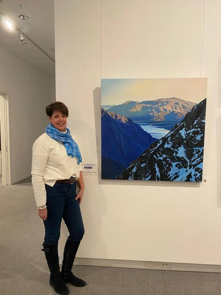 A woman standing next to a landscape painting of mountainous terrain and a lake.