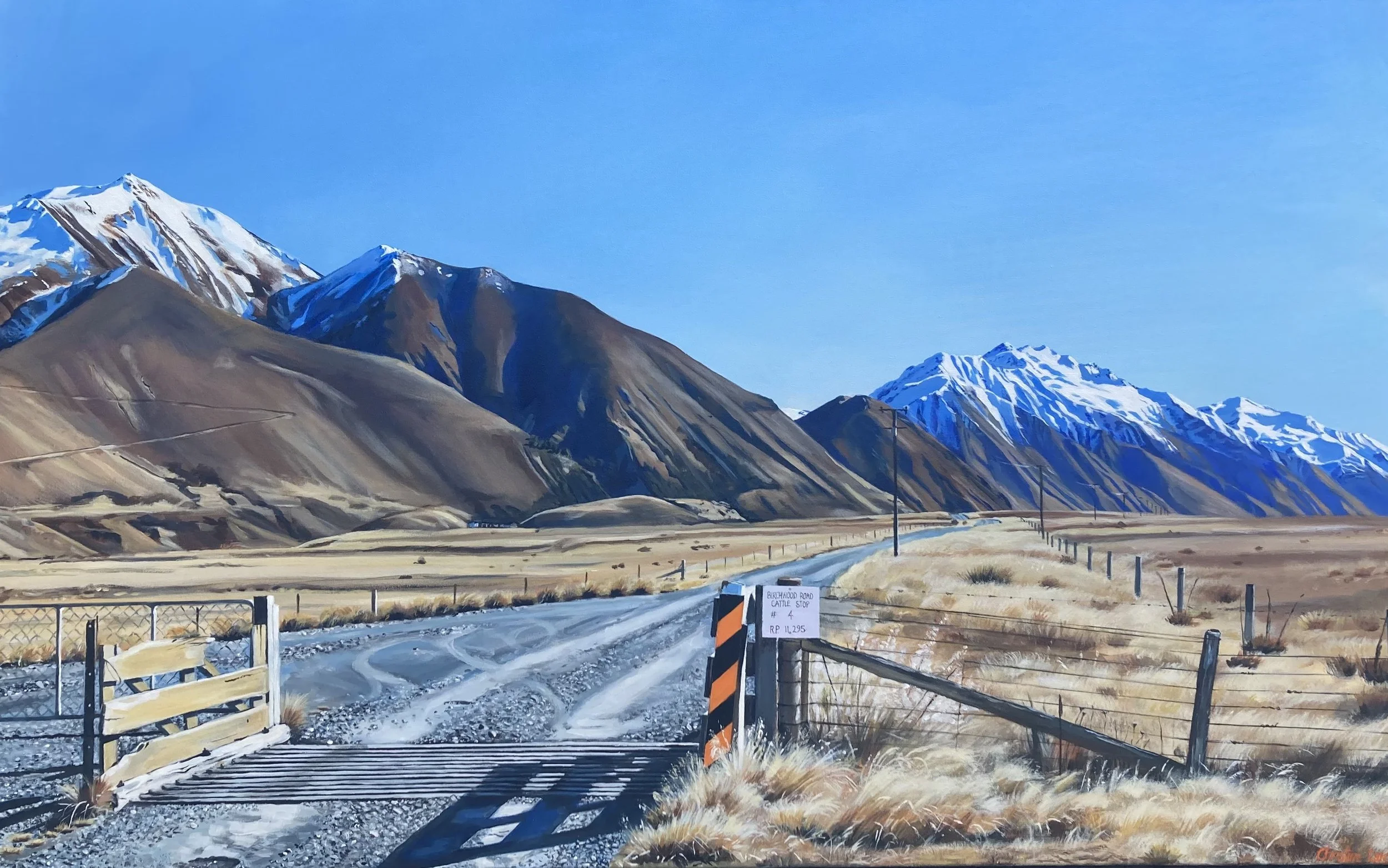 A rural landscape with snow-capped mountains in the background, a gravel road leading into the distance, and a wooden gate and fencing along the sides of the road under a clear blue sky.
