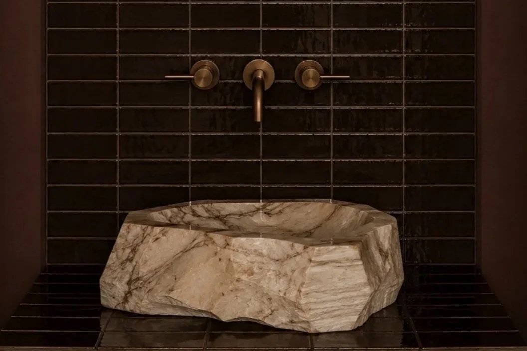 Marble stone sink mounted at the bathroom wall with black tiles and bronze faucet.