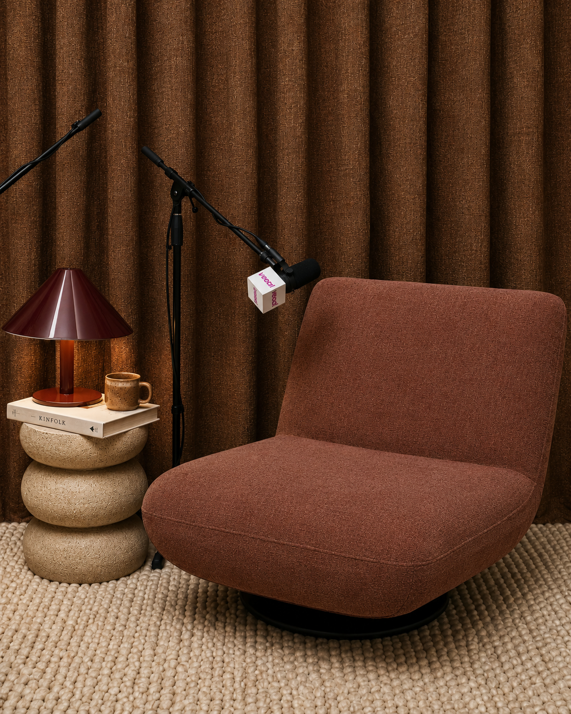 A cozy discussion or interview setup featuring a rust-colored upholstered chair, a small textured beige side table with a lamp, a book, and a coffee mug, and a microphone on a stand positioned in front of the chair, with a brown curtain backdrop.