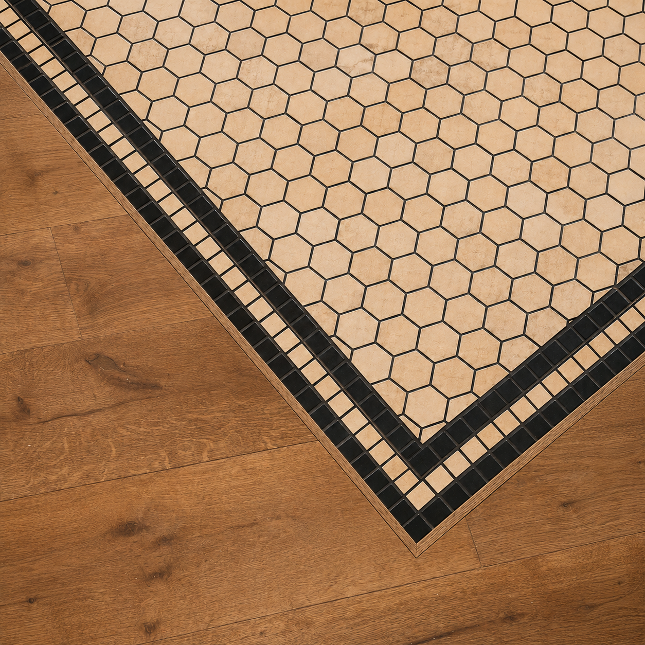 Corner of a wood floor meeting a tiled floor with a black border, hexagonal beige tiles, and small black square tiles.