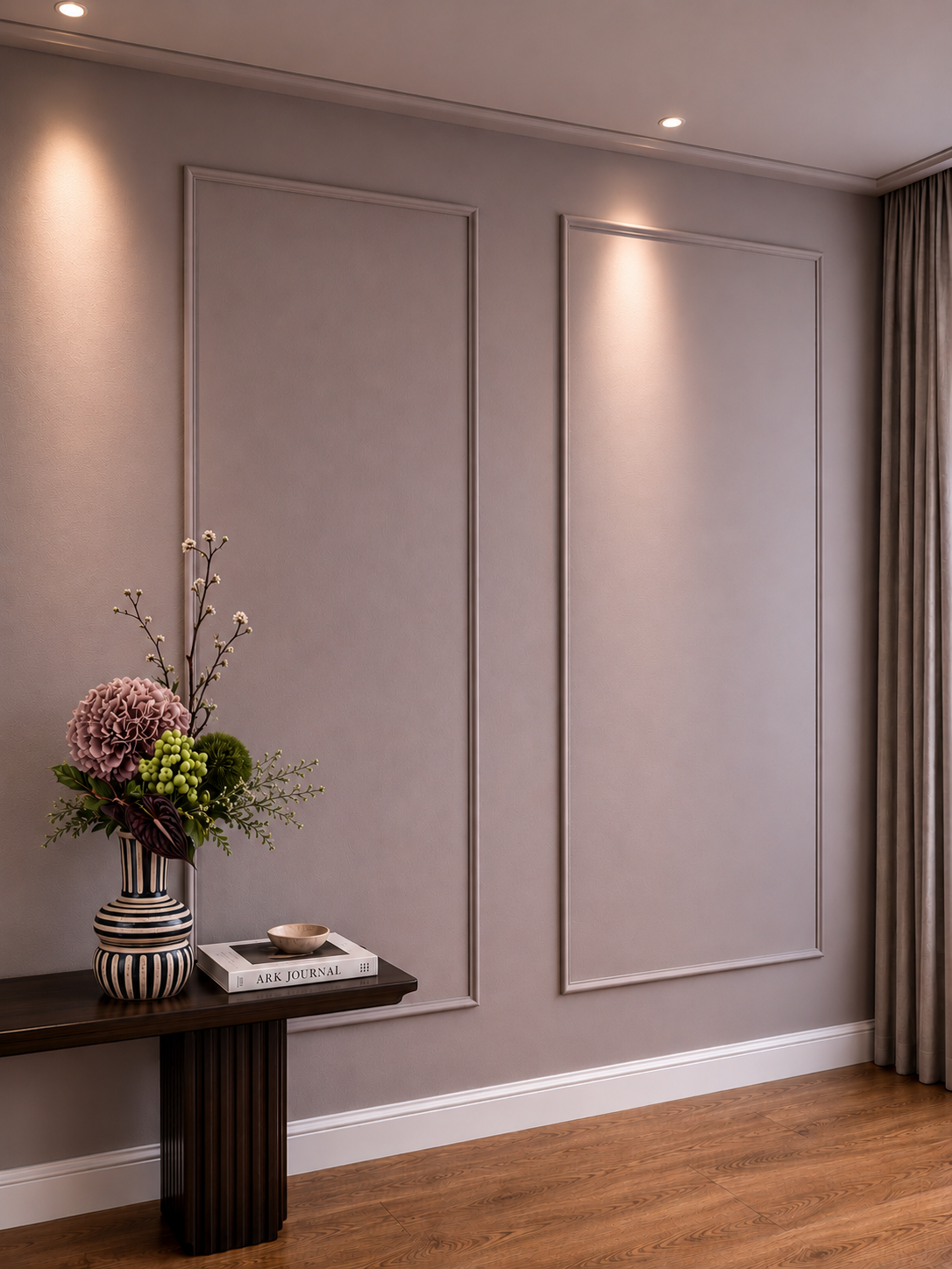 A minimalist interior wall with decorative molding, illuminated by spotlights, with a dark wood console table holding a floral arrangement in a black and white striped vase, a book titled 'ARK JOURNAL', and a small beige bowl. There are beige curtain