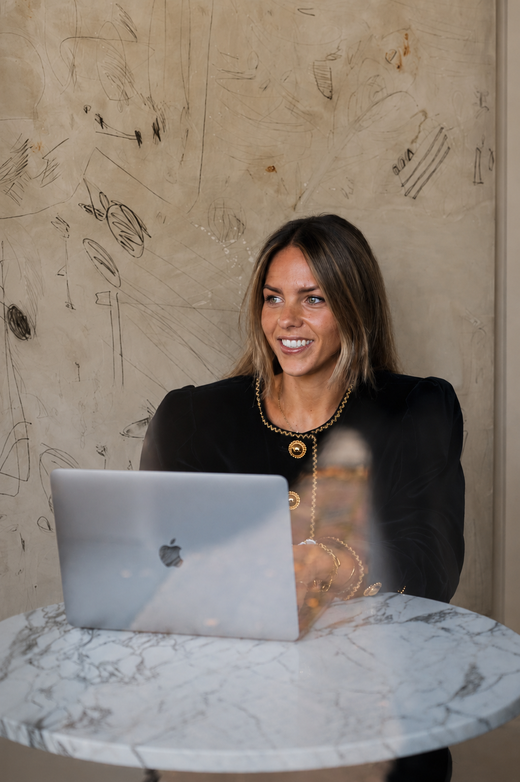 A woman with shoulder-length brown hair, wearing a black blazer with gold buttons and jewelry, sitting at a marble table with a silver MacBook in front of her, smiling and looking to her left, with a drawing-covered wall in the background.