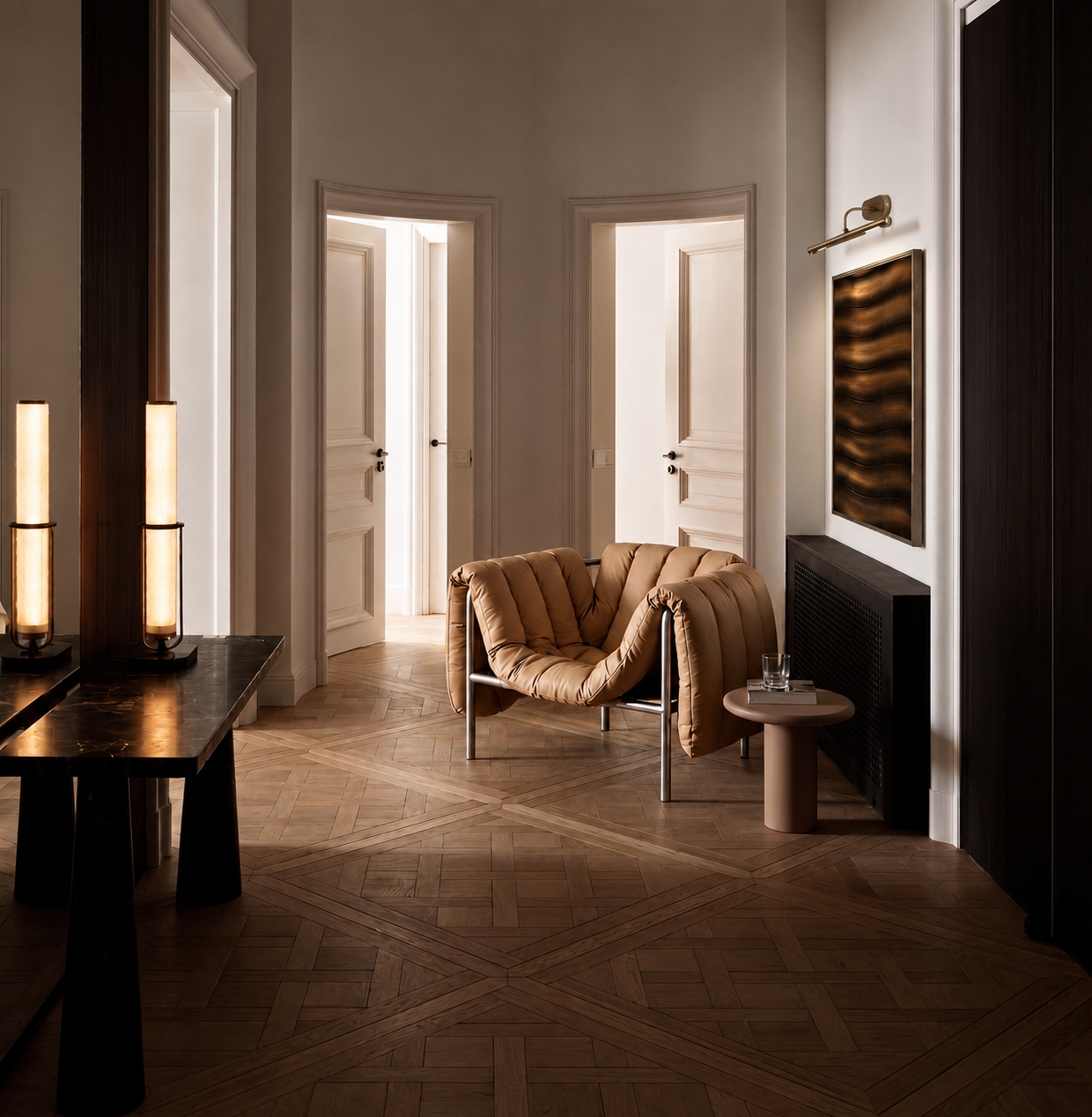A modern interior space with wooden flooring, beige walls, and white doors. There is a tan upholstered chair and a small round side table with a glass of water. A black vent and artwork are on the wall, with tall cylindrical lamps on a dark surface on the left side.