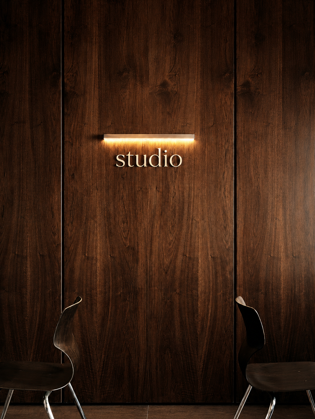 Wood-paneled wall with illuminated 'studio' sign and two black chairs in front.