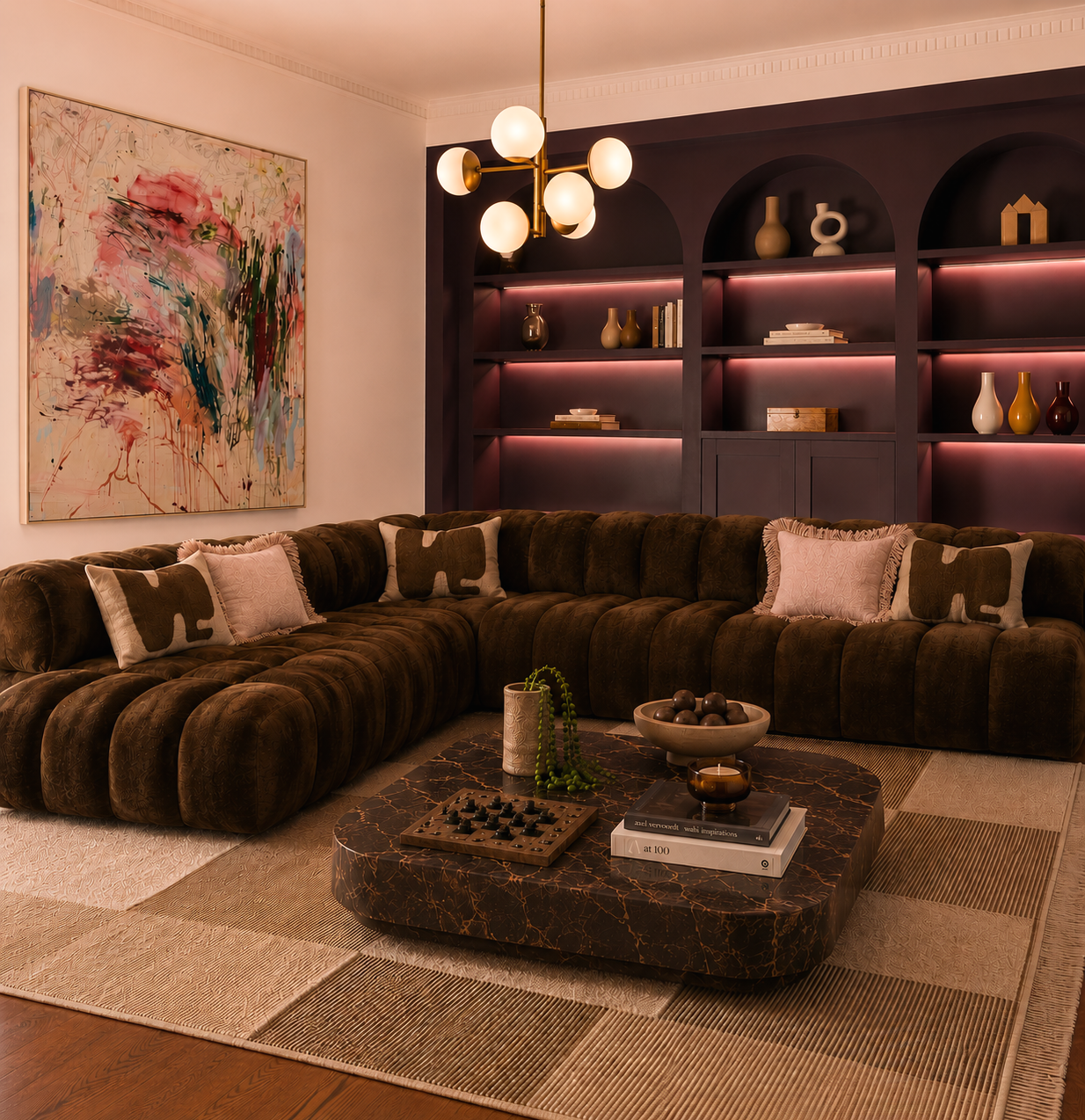 Living room with a large brown sectional sofa, abstract art on the wall, and a dark built-in bookshelf with decorative objects and books, illuminated by a modern chandelier.