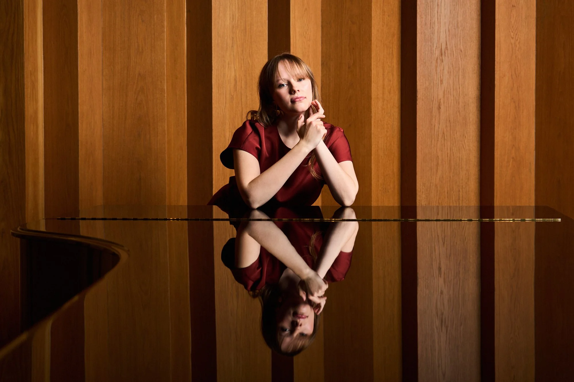 A woman with red hair in a maroon dress, sitting at a reflection piano with wooden panel background, resting her chin on her hand, looking thoughtfully.