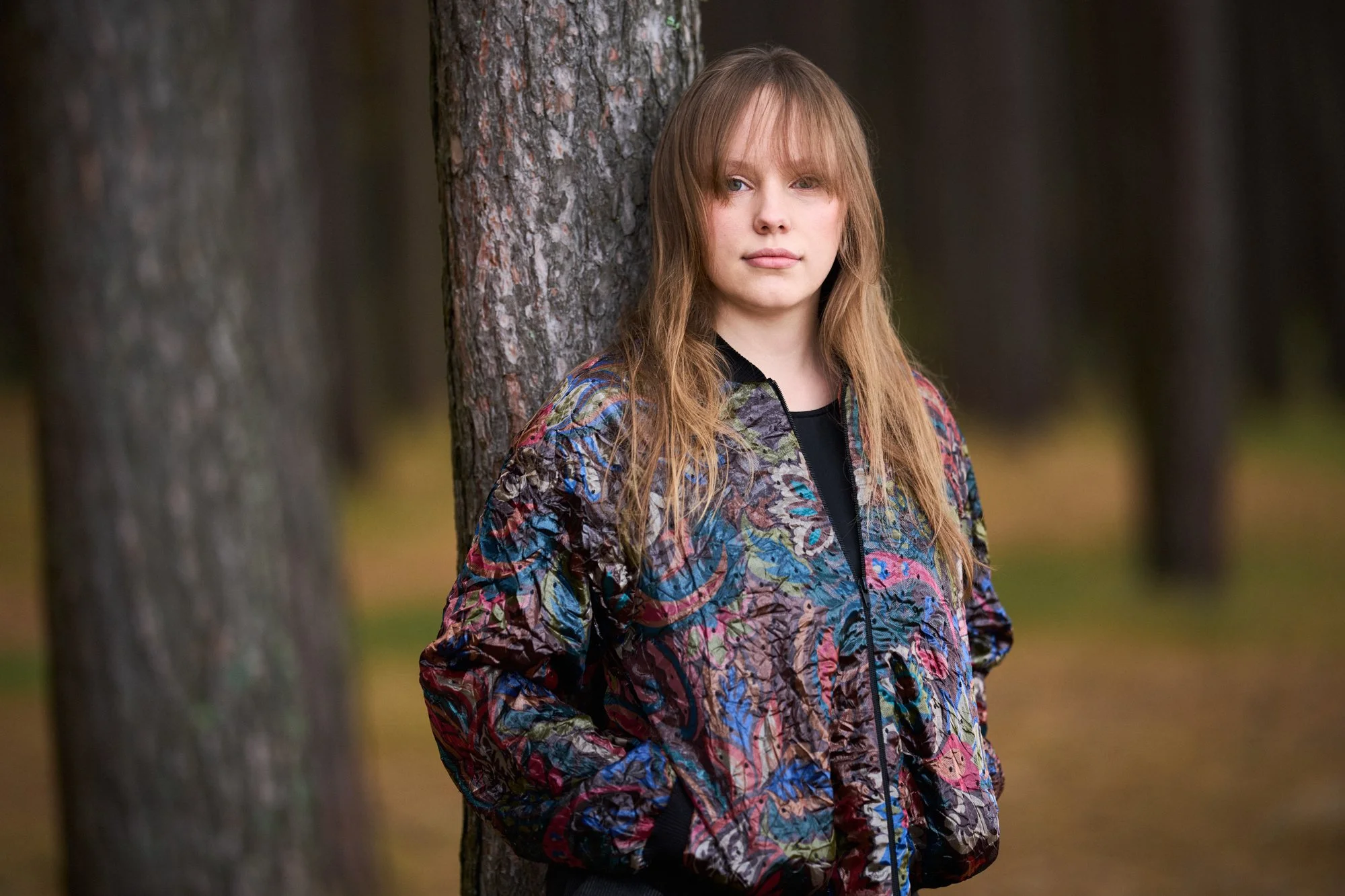 A young woman with light skin and long reddish-brown hair, wearing a colorful patterned jacket, leaning against a tree in a forest.
