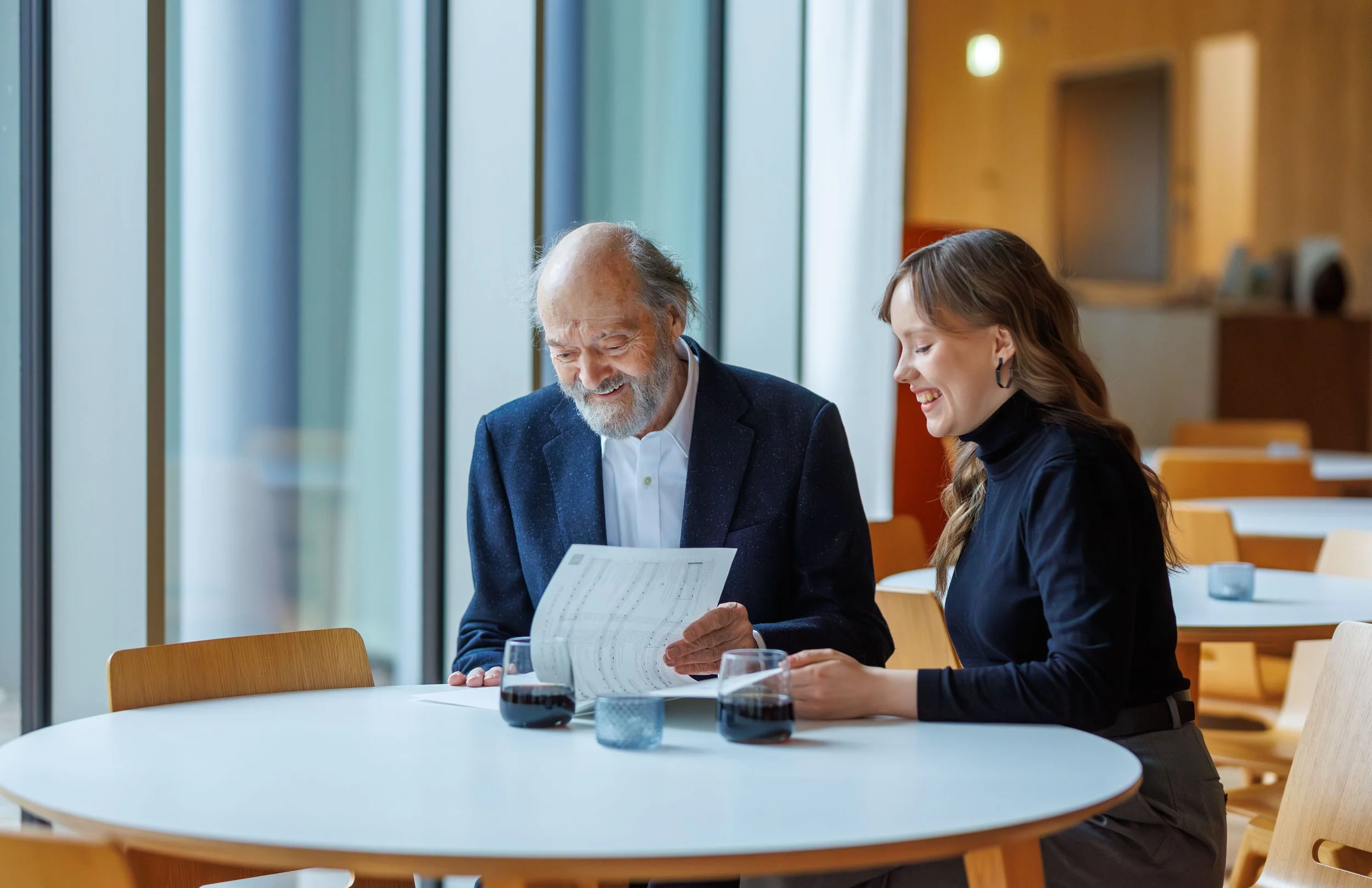 An elderly man and a young woman sit at a round table in a modern, well-lit cafe or restaurant. They are smiling and looking at a paper or menu the man is holding. There are glasses of red wine and small blue candles on the table, and large windows w