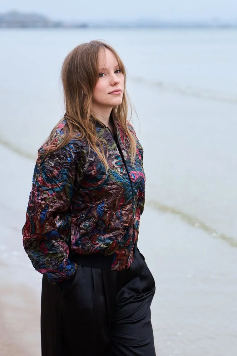 A young woman with long, light brown hair standing on a beach near the water, wearing a colorful patterned jacket and black pants.