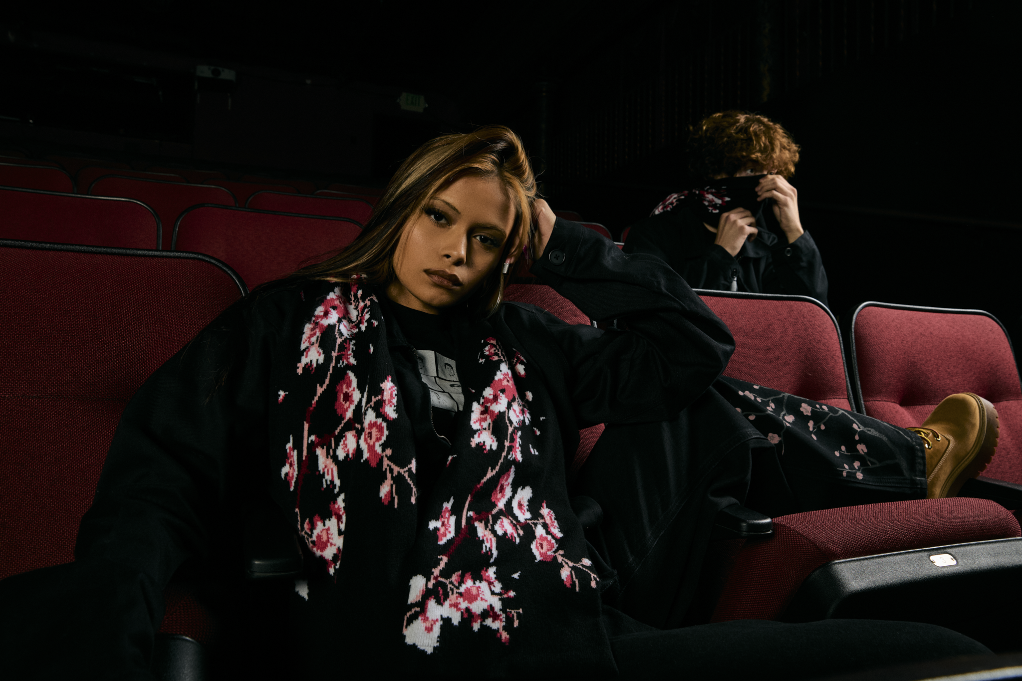 A young woman with long brown hair, lying on red theater seats, wearing a black jacket with pink and white floral patterns, and yellow boots. She is resting her head on her hand and looking at the camera. In the background, a young man with curly red hair is sitting, holding a mask up to his face, with only part of his face visible.