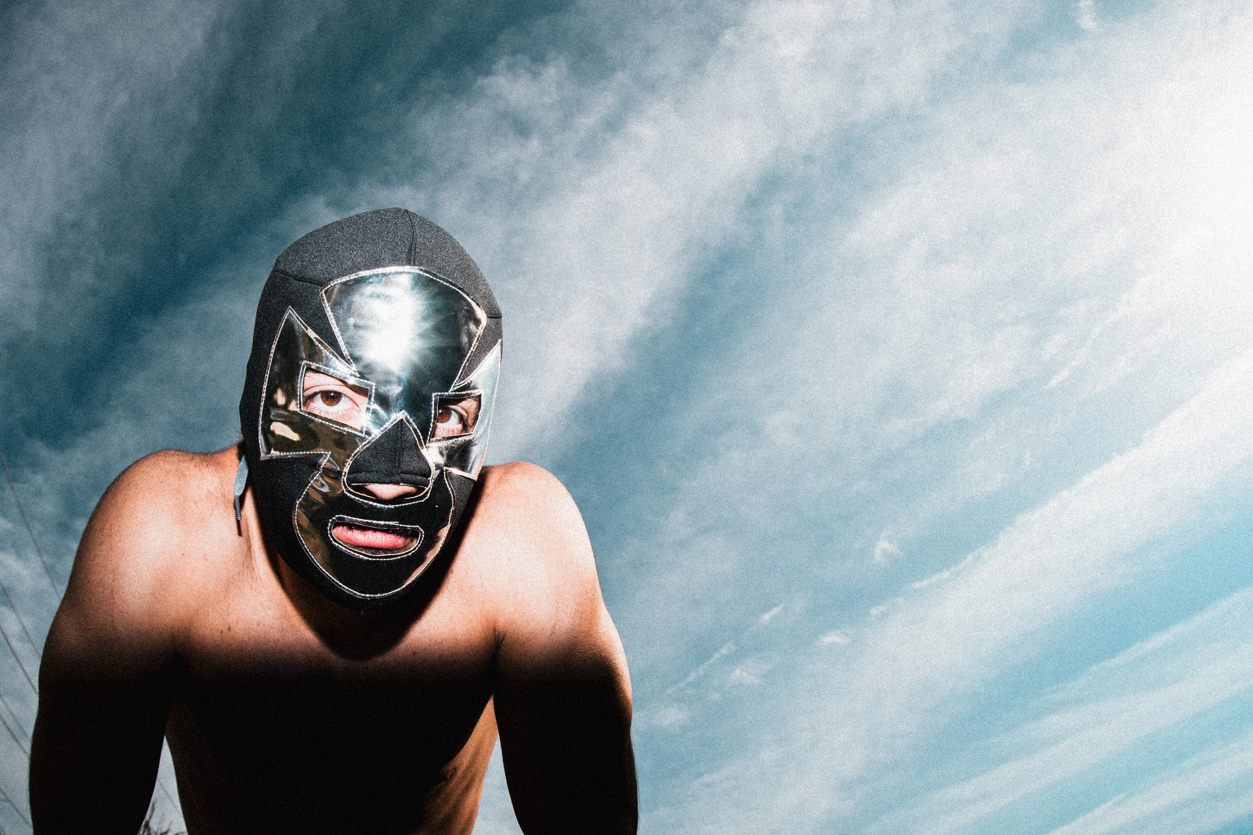 Person wearing a black and silver luchador mask with brown eyes, squatting shirtless against a cloudy sky.