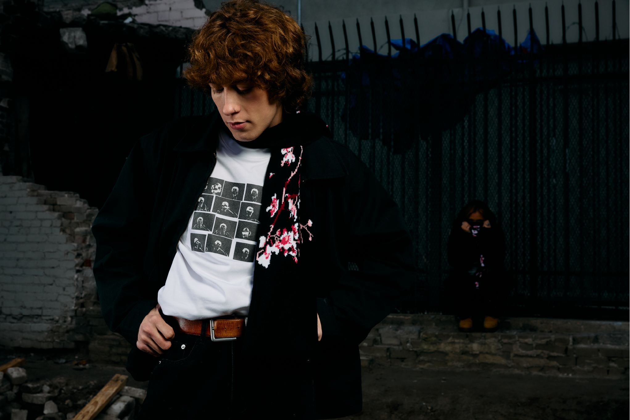 Young man with curly hair in a black jacket and graphic T-shirt, standing outdoors in an urban area with construction debris and a fence in the background.
