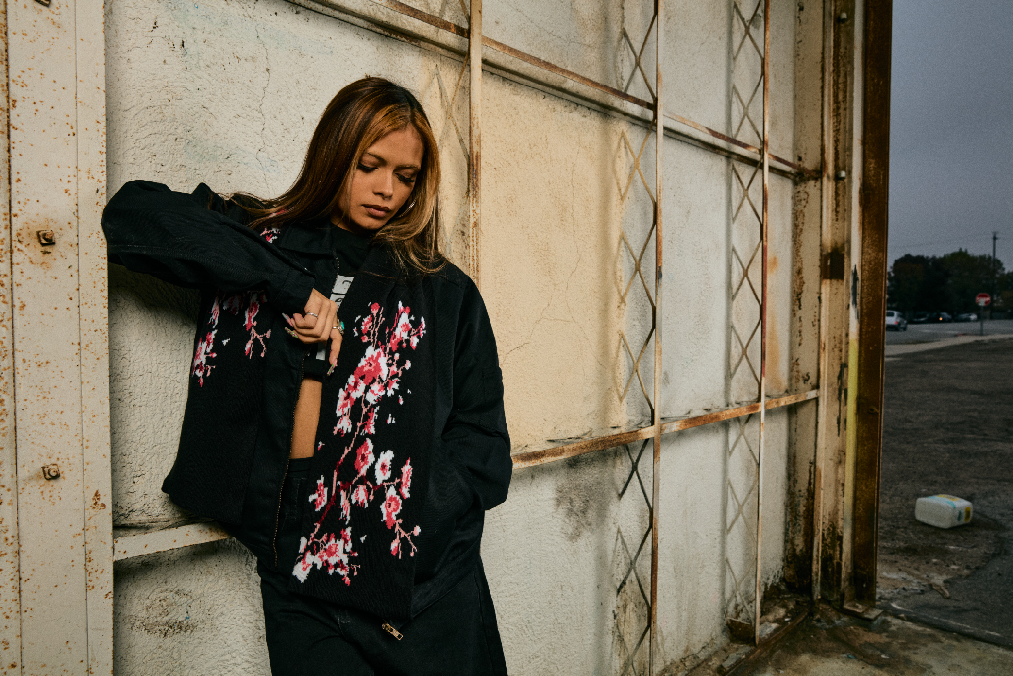 A young woman with long hair is leaning against a rusty metal structure attached to a weathered wall. She is wearing a black jacket with floral embroidery, her eyes are closed, and she appears to be in a contemplative mood. The background includes an