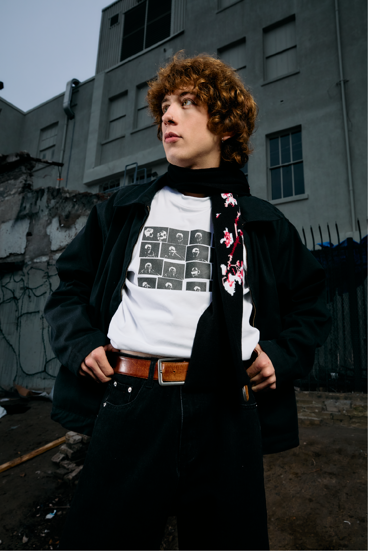 A person with curly brown hair, wearing a black jacket, a white graphic t-shirt, and a brown belt, stands outside against a gray industrial building with a cloudy sky.