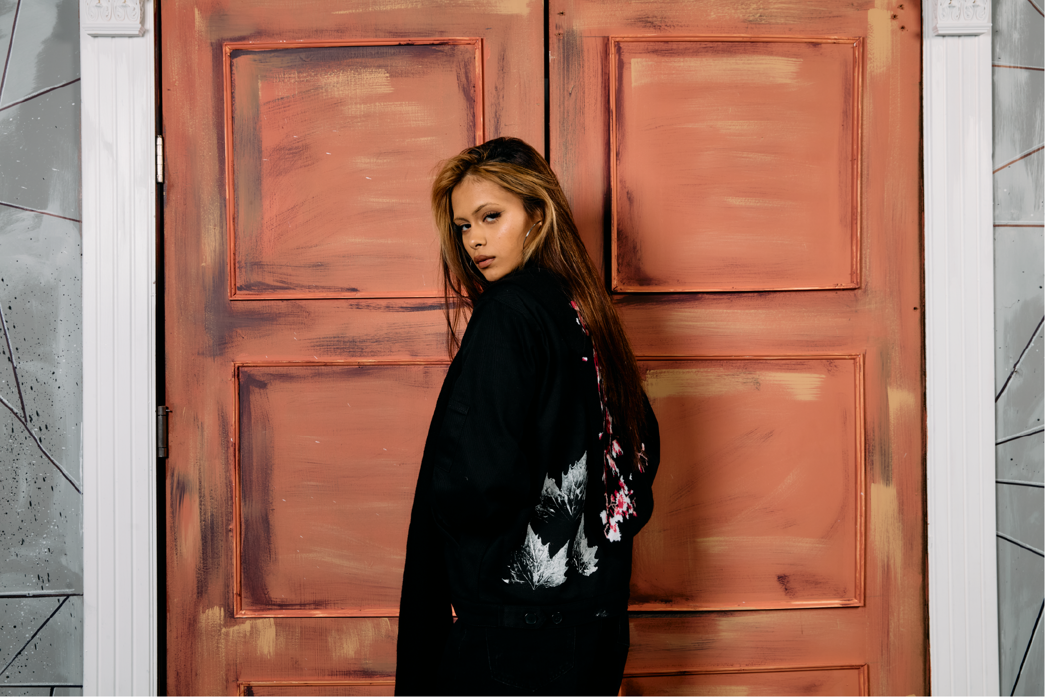 Young woman with long hair standing in front of a red door, wearing a black jacket with a white and pink floral design on the back.