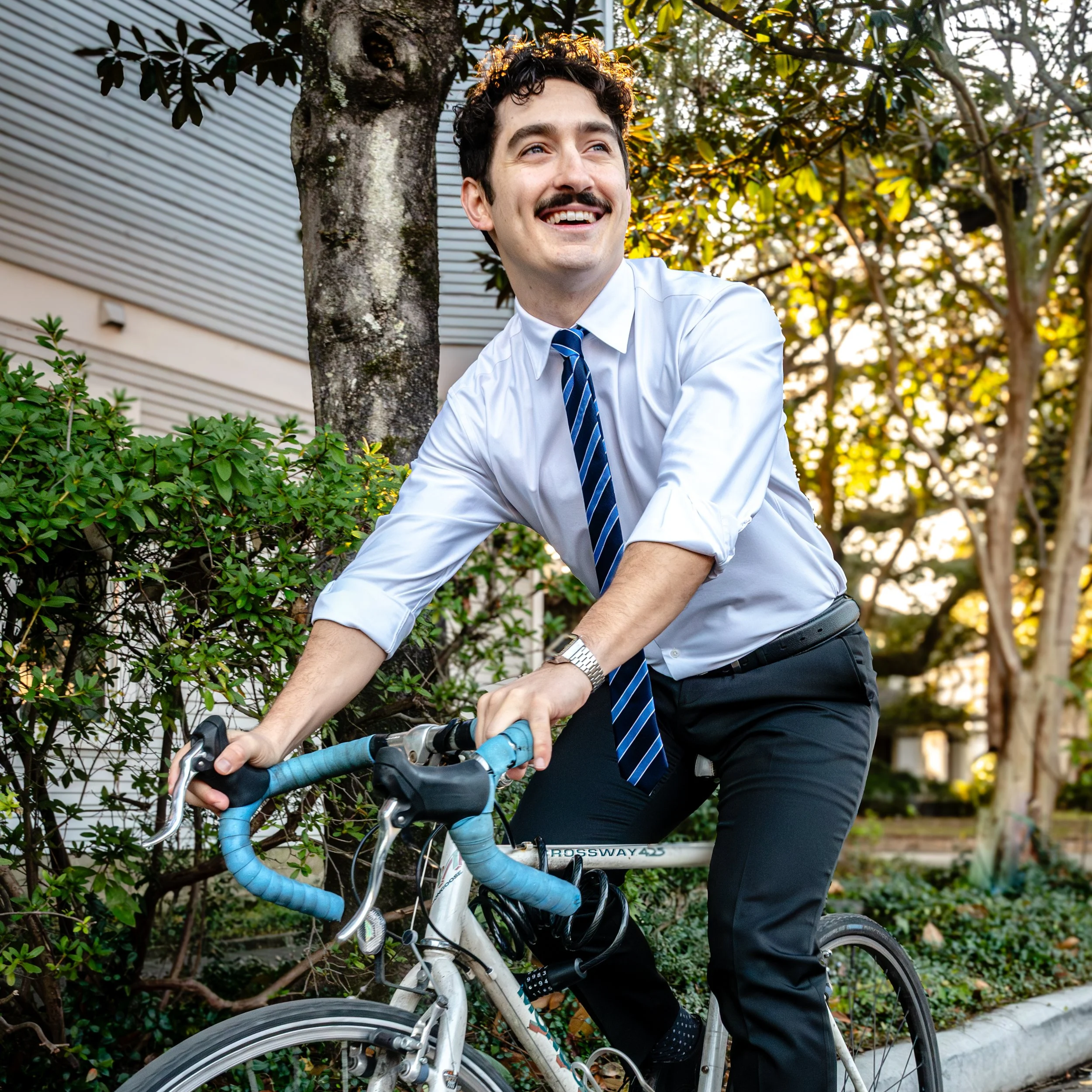 Joe Panzarella wearing a white dress shirt, blue tie, and black pants, riding a bicycle outdoors with trees and greenery in the background.
