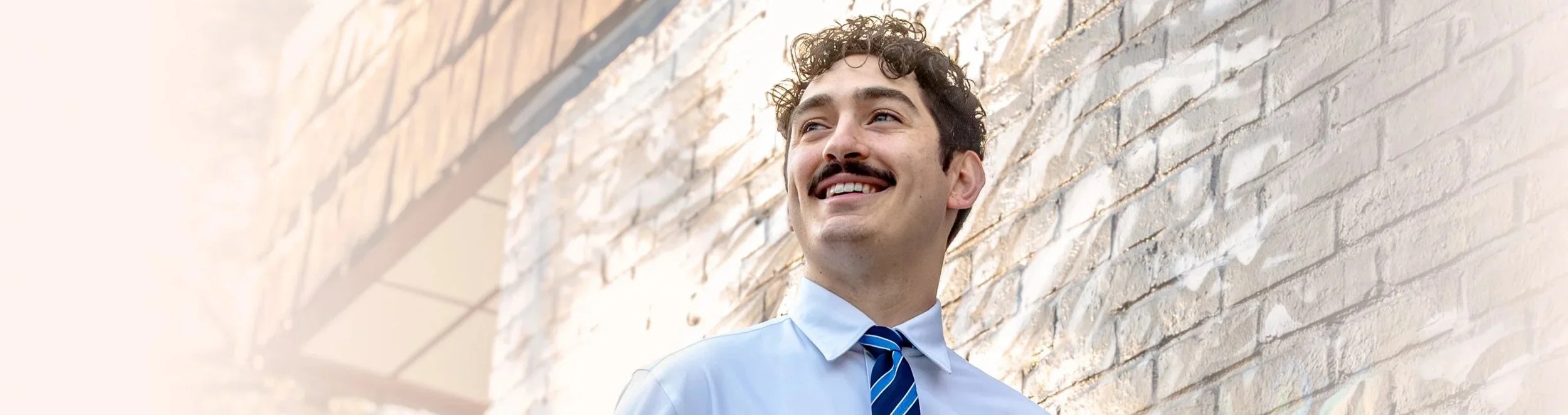 A man with a mustache in business attire standing against a brick wall, looking to his left with a cheerful expression.