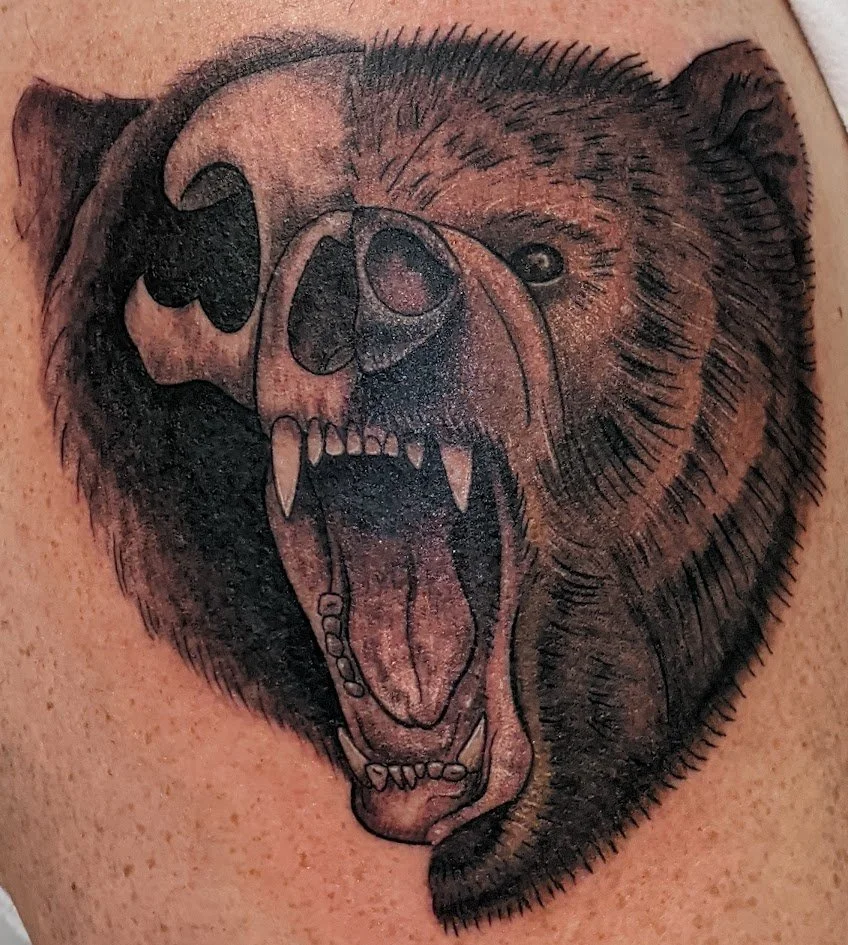 Tattoo of a bear with its mouth open, showing sharp teeth, combining the face of a bear and a skull in black ink.