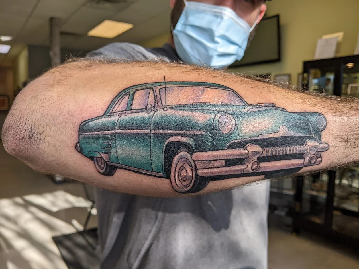 Tattoo of a vintage blue-green car on a person's upper arm, with a person wearing a face mask in the background.